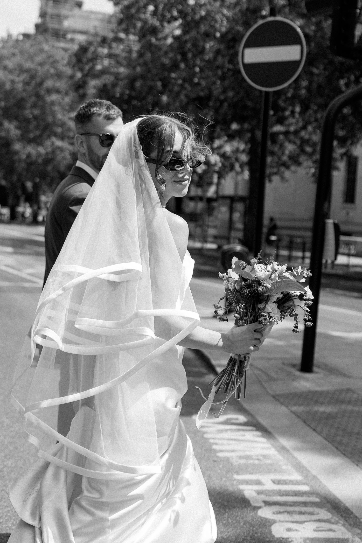 Smiling bride in veil and sunglasses crossing street with groom.