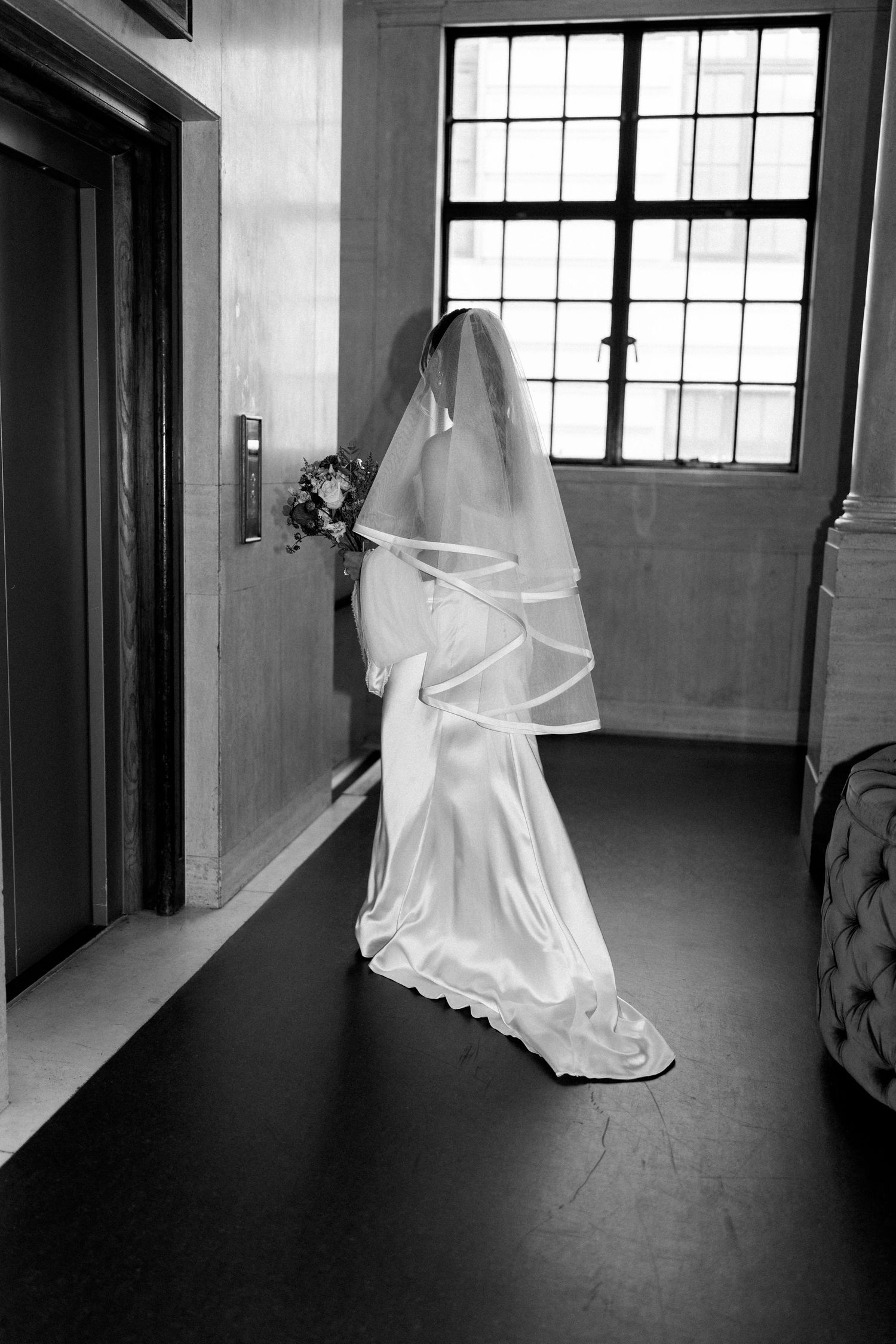 Bride in white wedding gown and veil holding bouquet waiting by an elevator.
