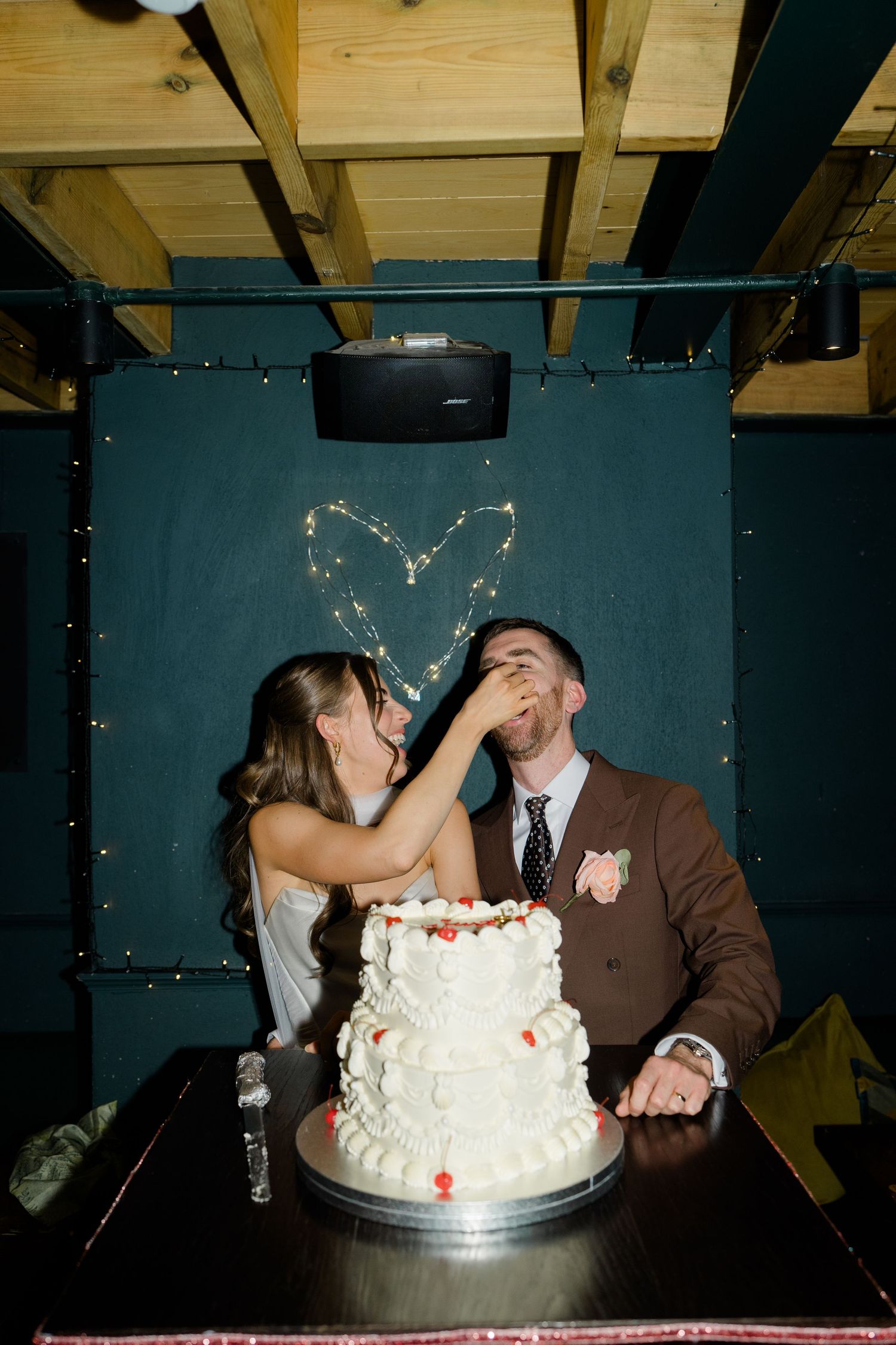 Bride feeding wedding cake to groom at reception table with white tiered cake and fairy light heart backdrop.
