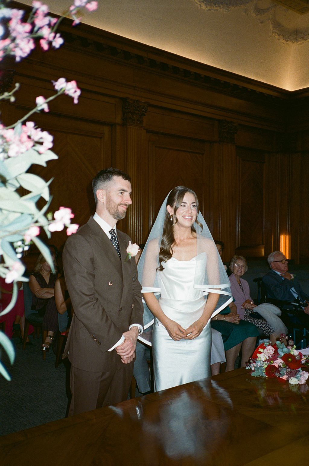 Bride and groom smiling at wedding ceremony in wood-paneled room with floral arrangements.