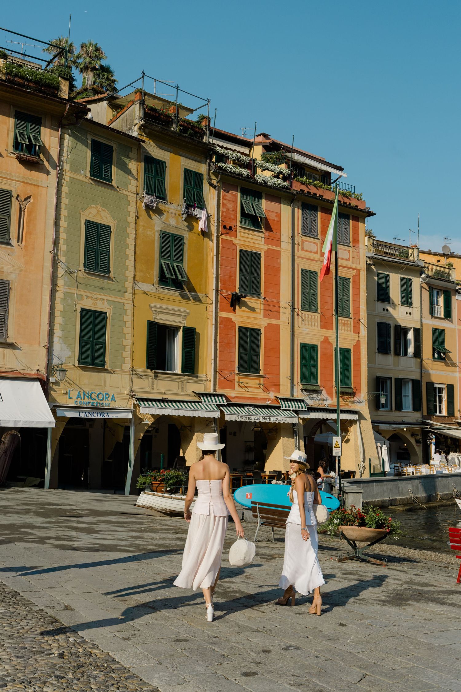 Two women in white dresses and hats walking past colorful Italian buildings in Portofino harbor.