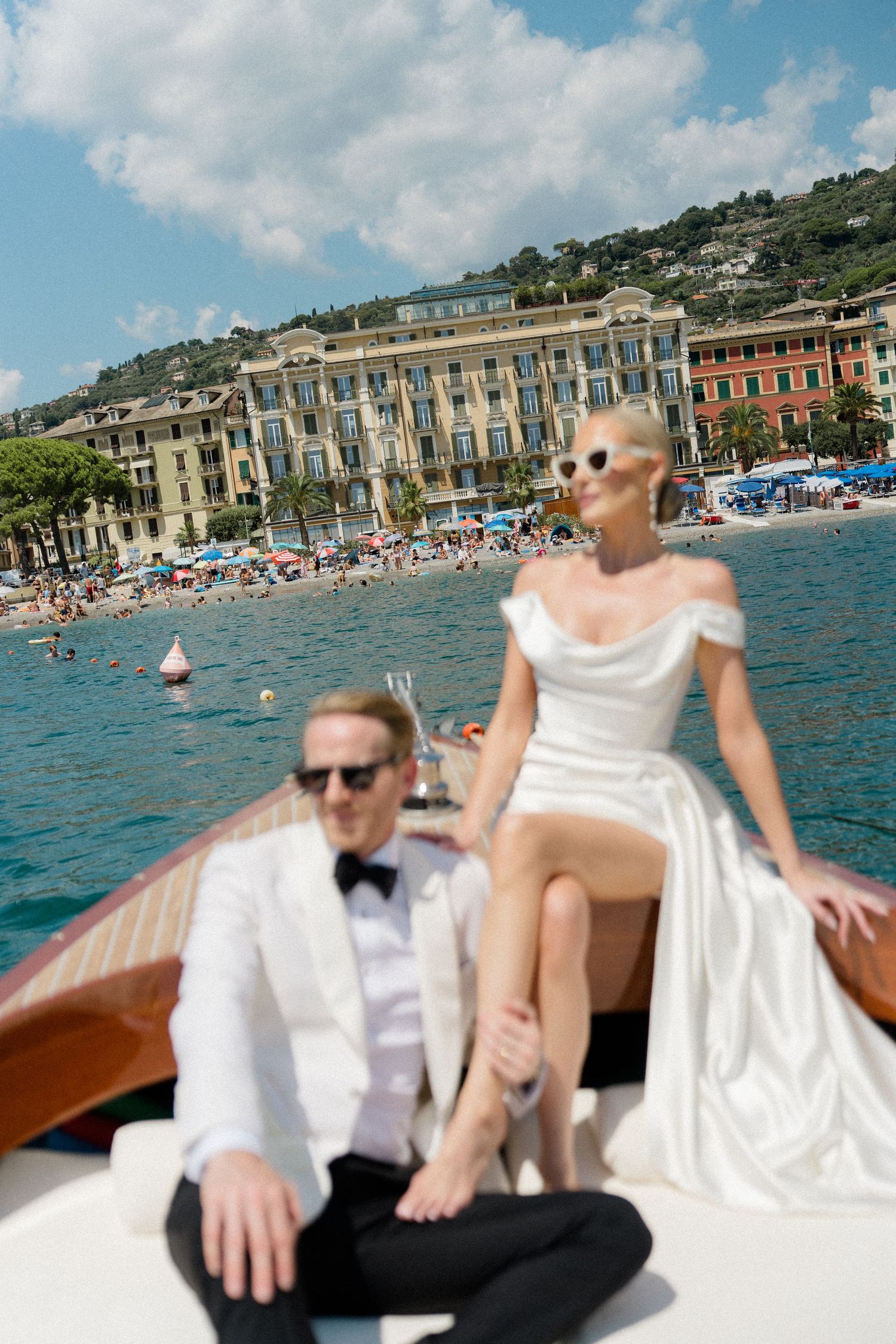 Bride in white off-shoulder gown and groom in tuxedo pose on boat near Italian coastal town.