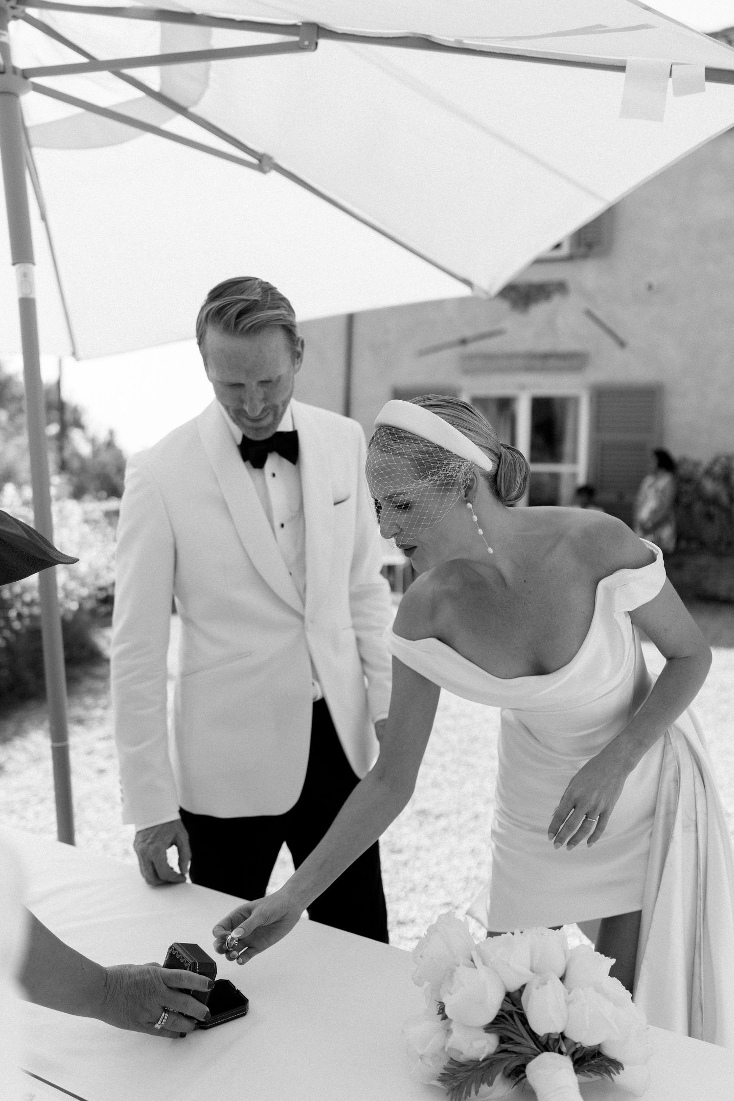 Black and white photo of a smiling bride signing wedding documents with groom at an outdoor ceremony table.