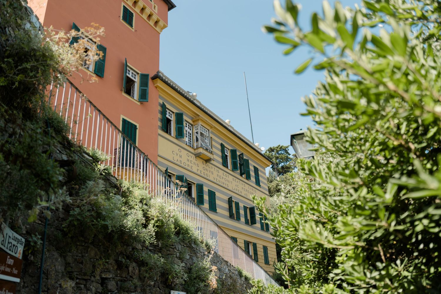 Colorful Italian coastal building with green shutters surrounded by lush Mediterranean vegetation.
