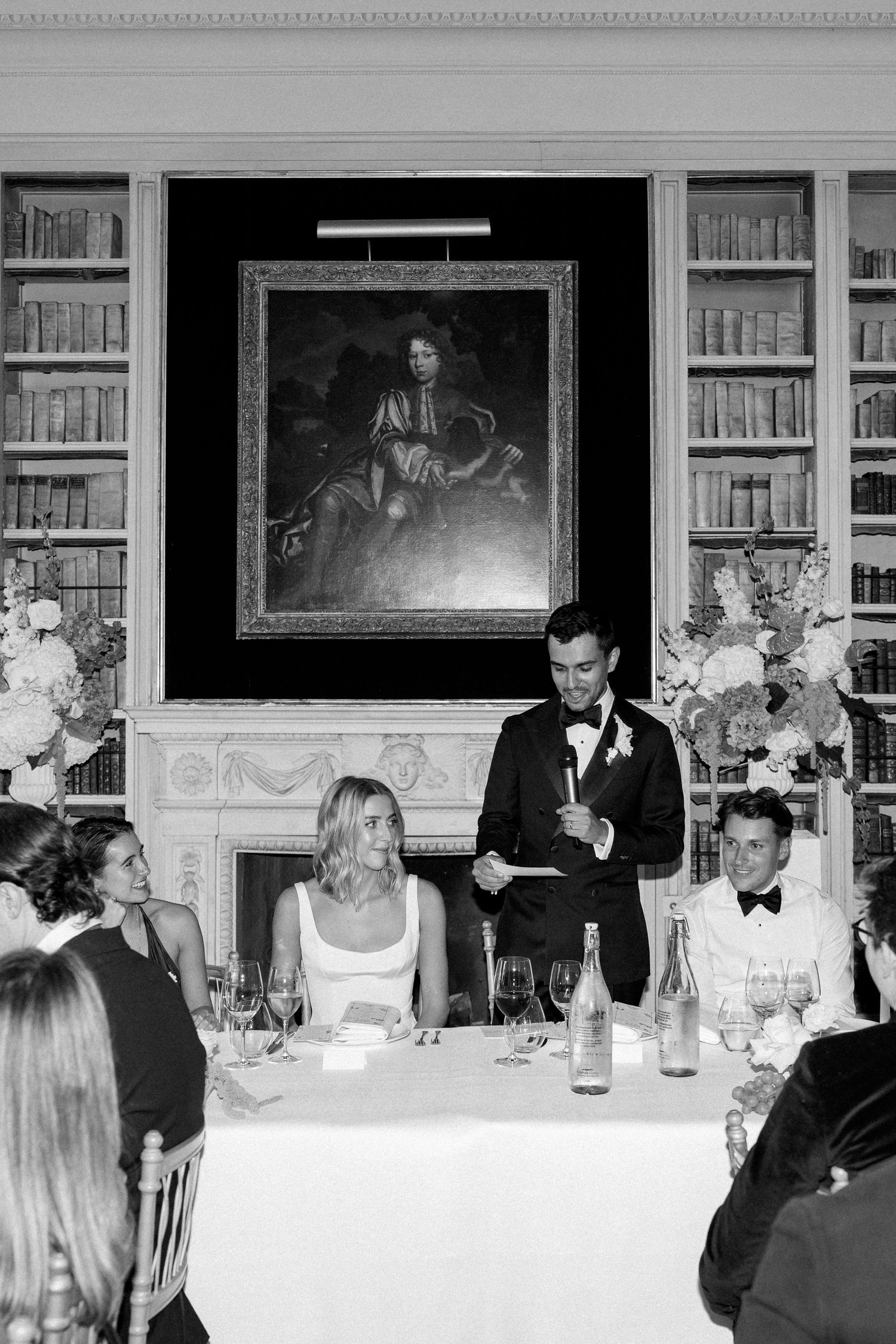 Groom delivering speech at wedding reception in a grand library with floral arrangements and portrait.