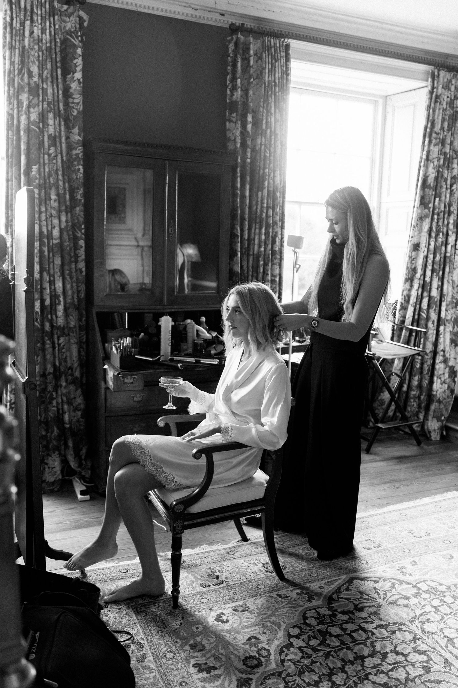 Black and white photo of a bride in a robe seated while a stylist prepares her hair in an elegant room.