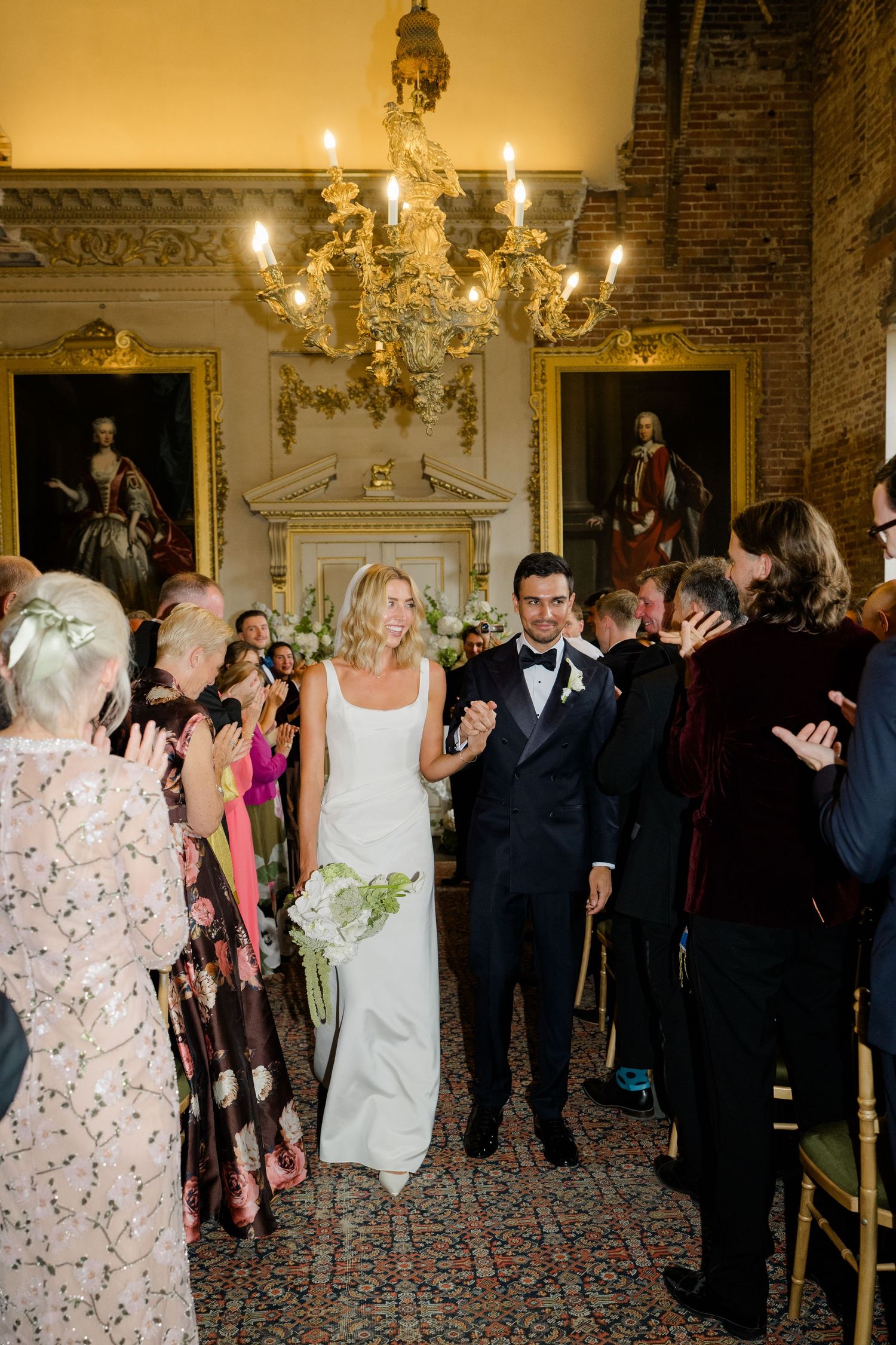 Bride and groom walk down the aisle as wedding guests applaud in an elegant room with gold chandelier.