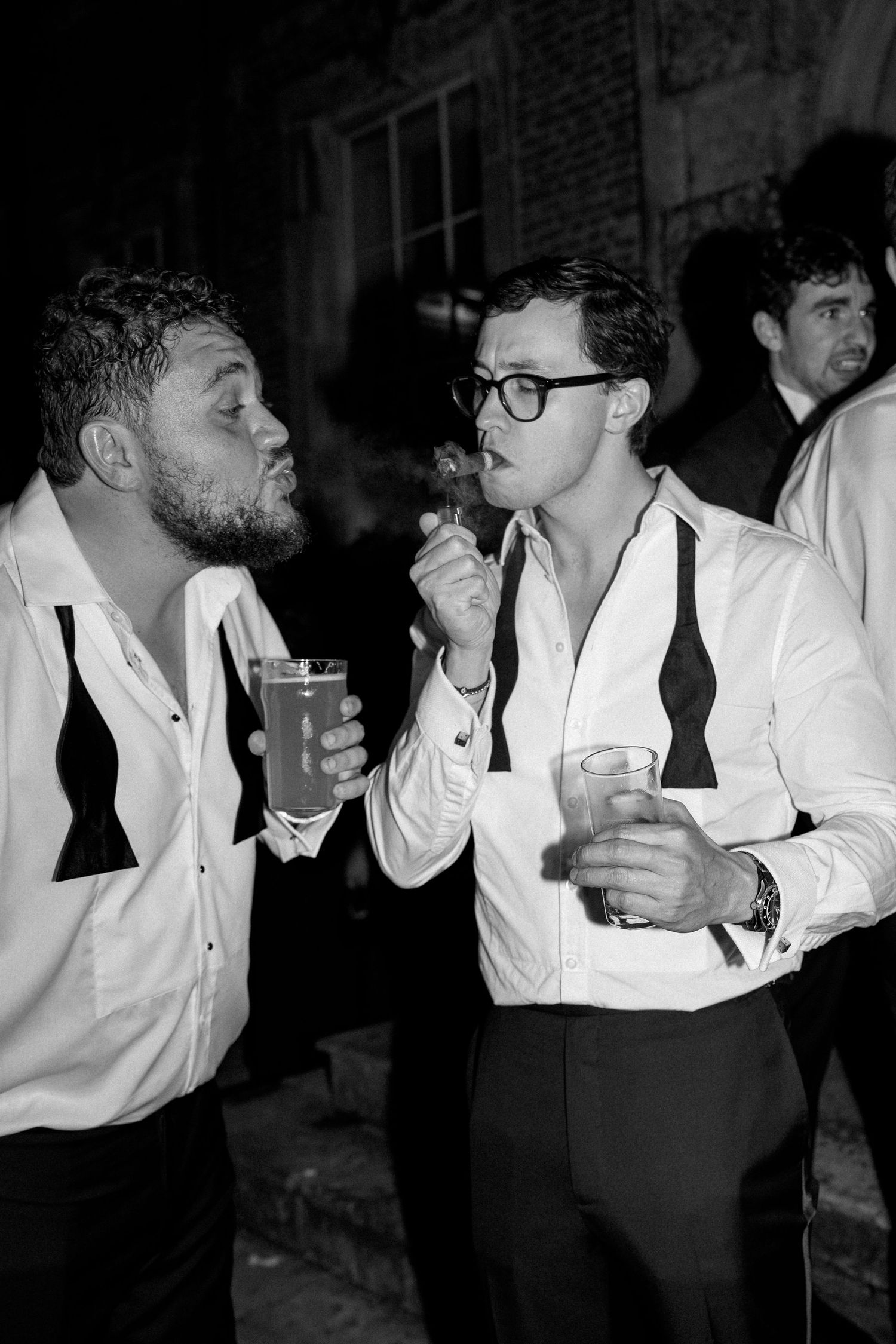 Two men in white shirts and bow ties sharing drinks and a cigar at a nighttime outdoor event.