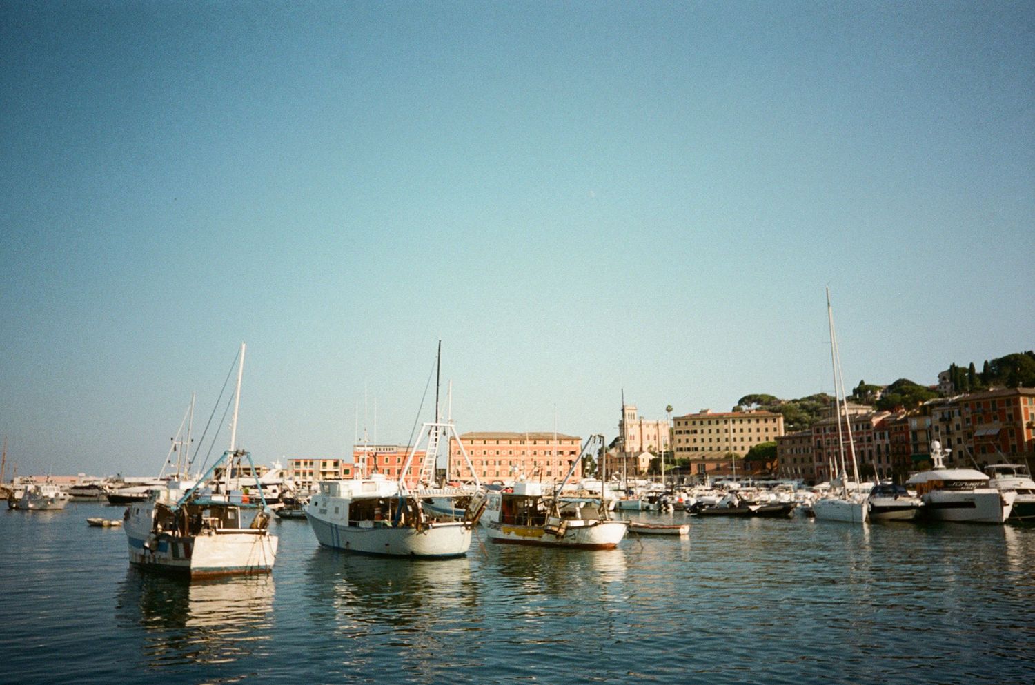 Sailboats and yachts moored in sunny Italian harbor with historic waterfront buildings backdrop.