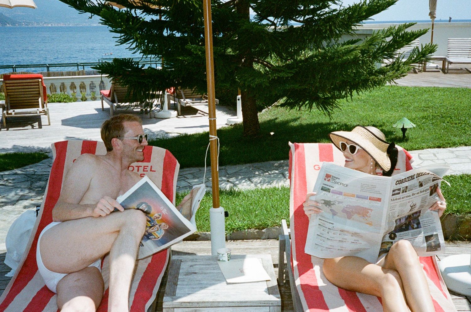 Couple relaxing on red and white striped sun loungers reading magazines by hotel poolside waterfront.