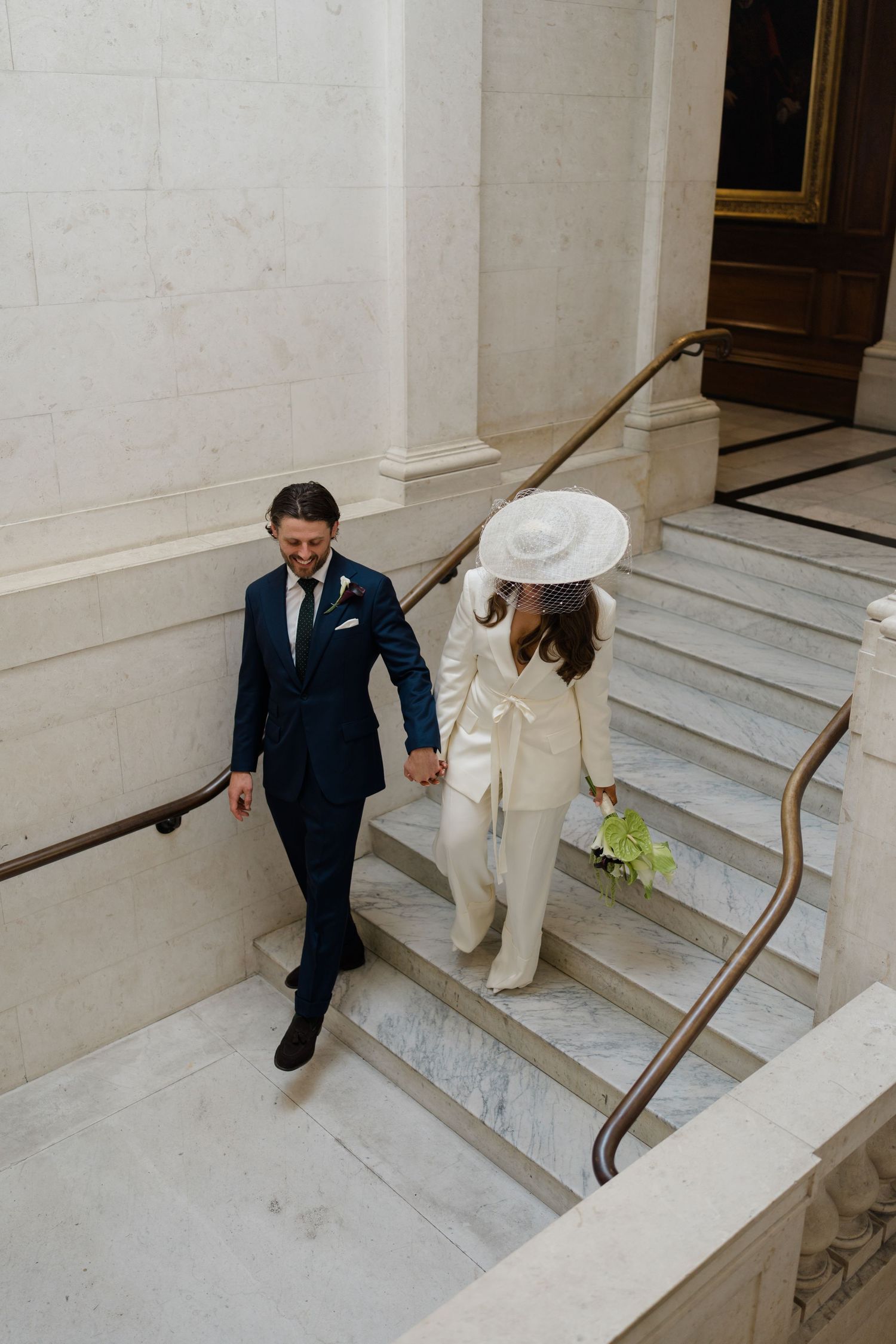 Bride in white jumpsuit and hat holds groom's hand walking up marble stairs at elegant wedding venue.