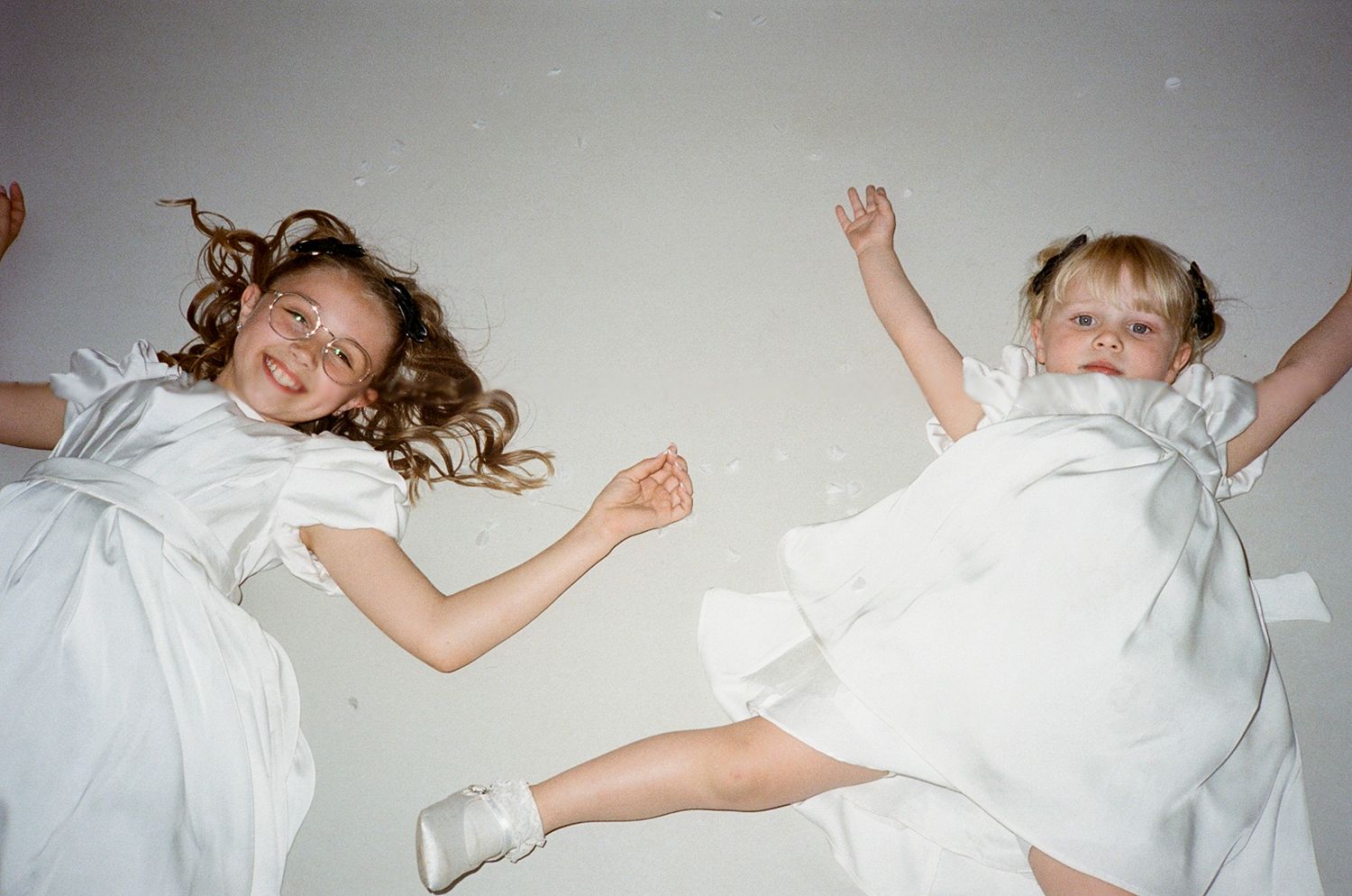 Two joyful girls in white dresses jumping mid-air with arms raised, captured from below against a light background.