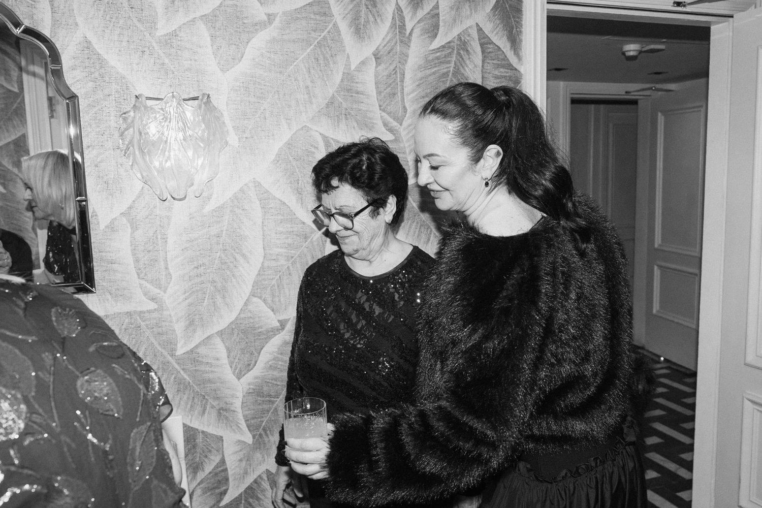 Two women in black outfits share a moment in a floral wallpapered room in black and white photo.