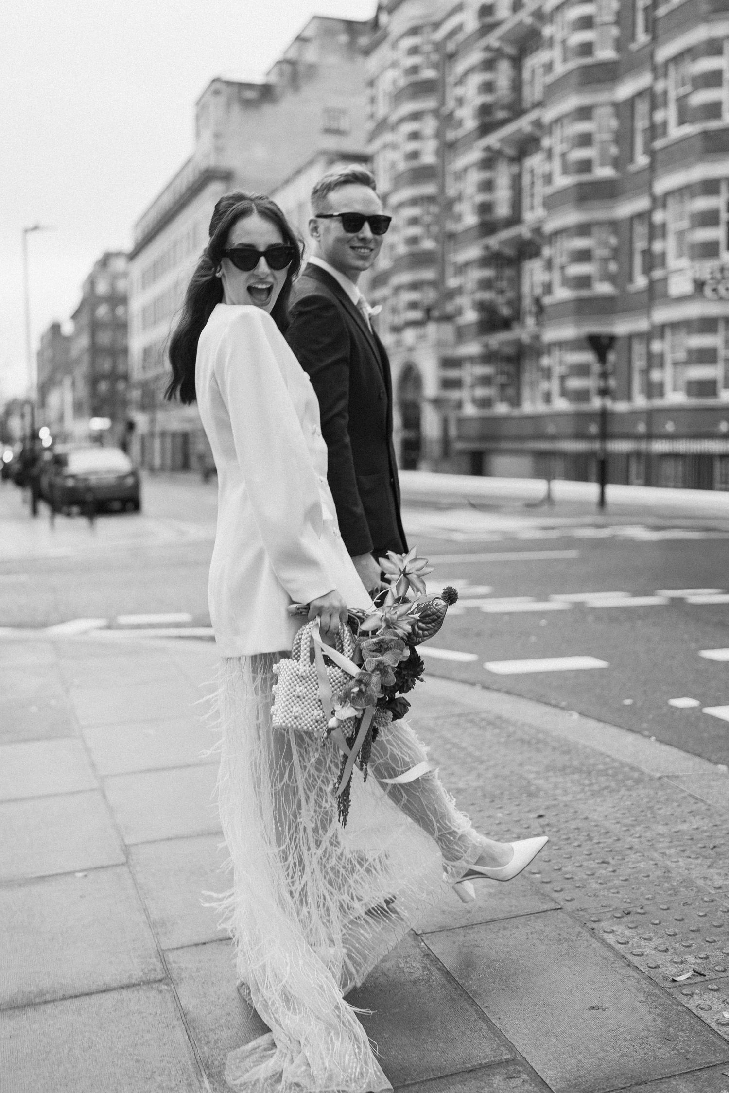 Black and white photo of stylish newlyweds walking London street, bride in feathered skirt and boots.