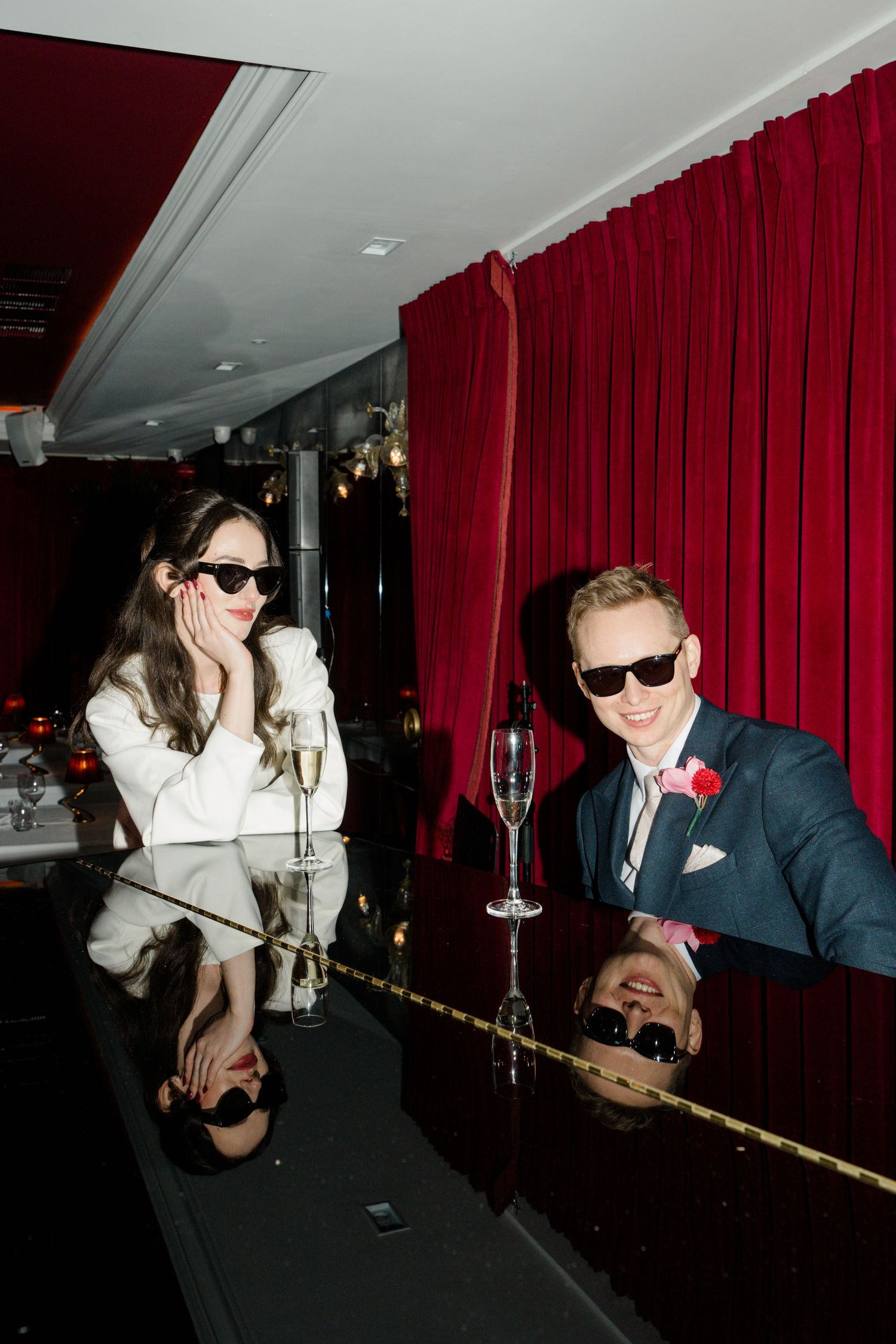 Bride and groom in sunglasses enjoy champagne at a mirrored bar with red velvet curtains at wedding reception.
