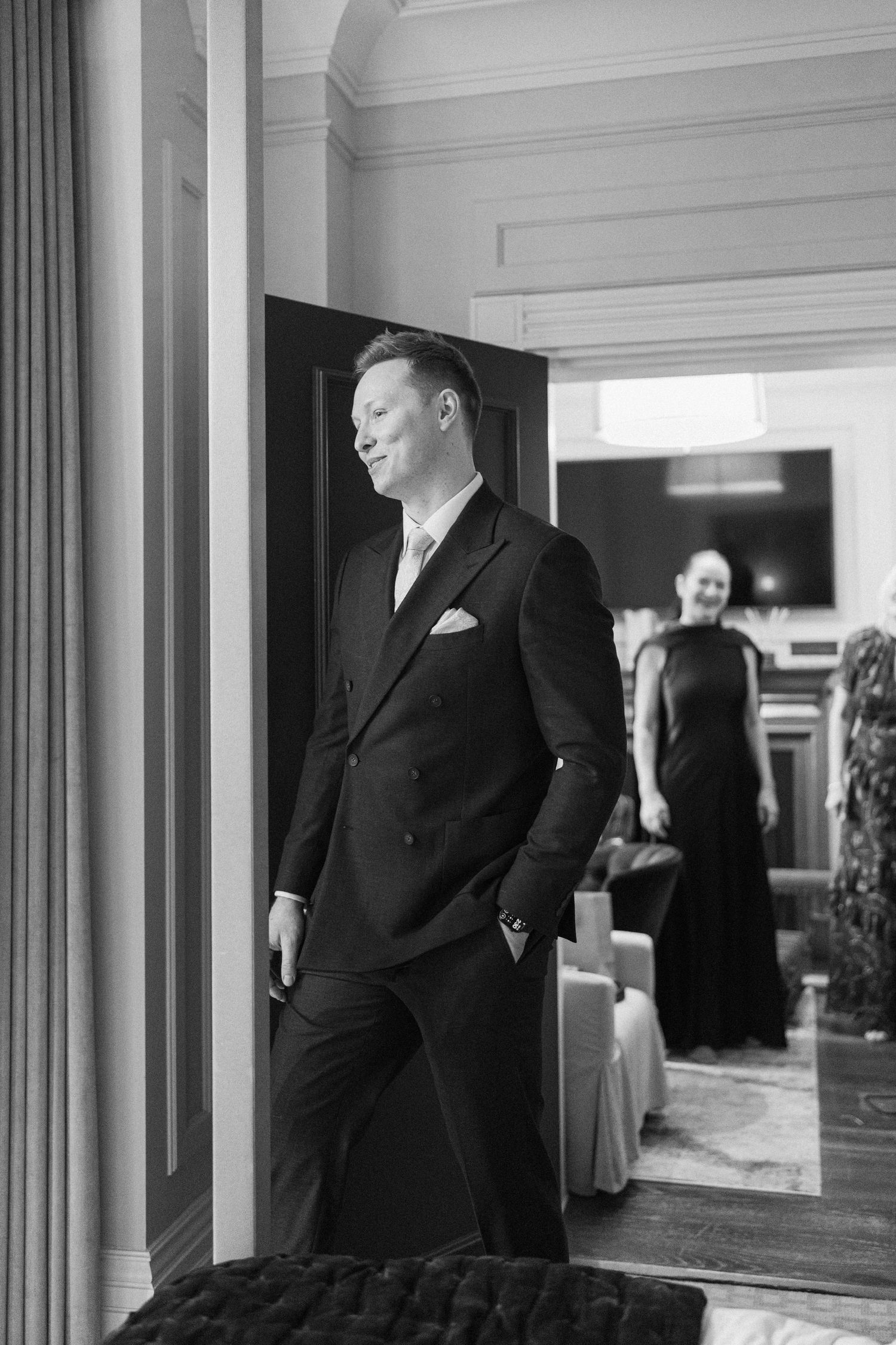 Groom in black double-breasted suit standing in doorway with woman in background, black and white.