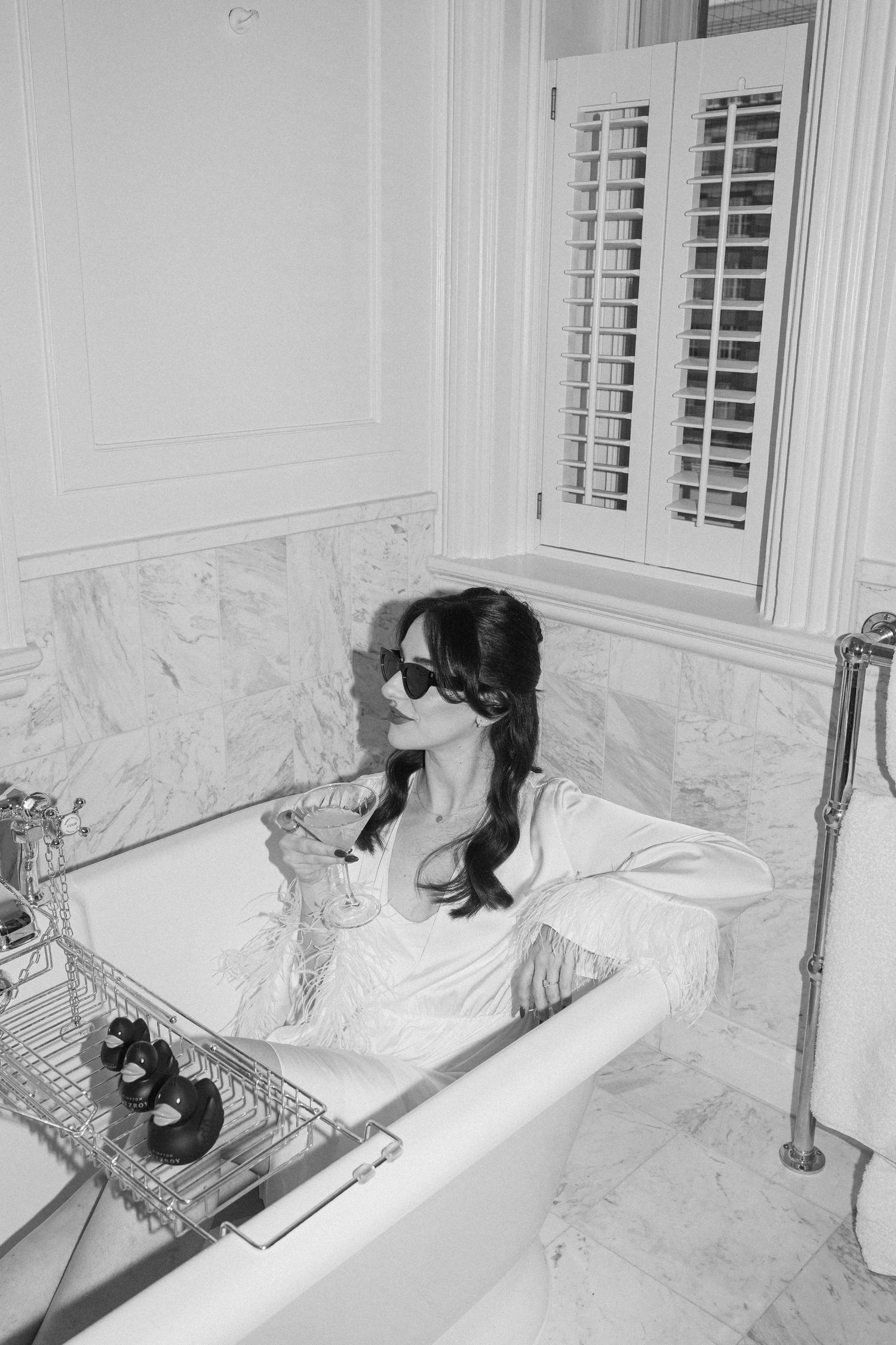 A glamorous bride in a white robe relaxes in a marble bathtub holding a champagne glass wearing sunglasses.