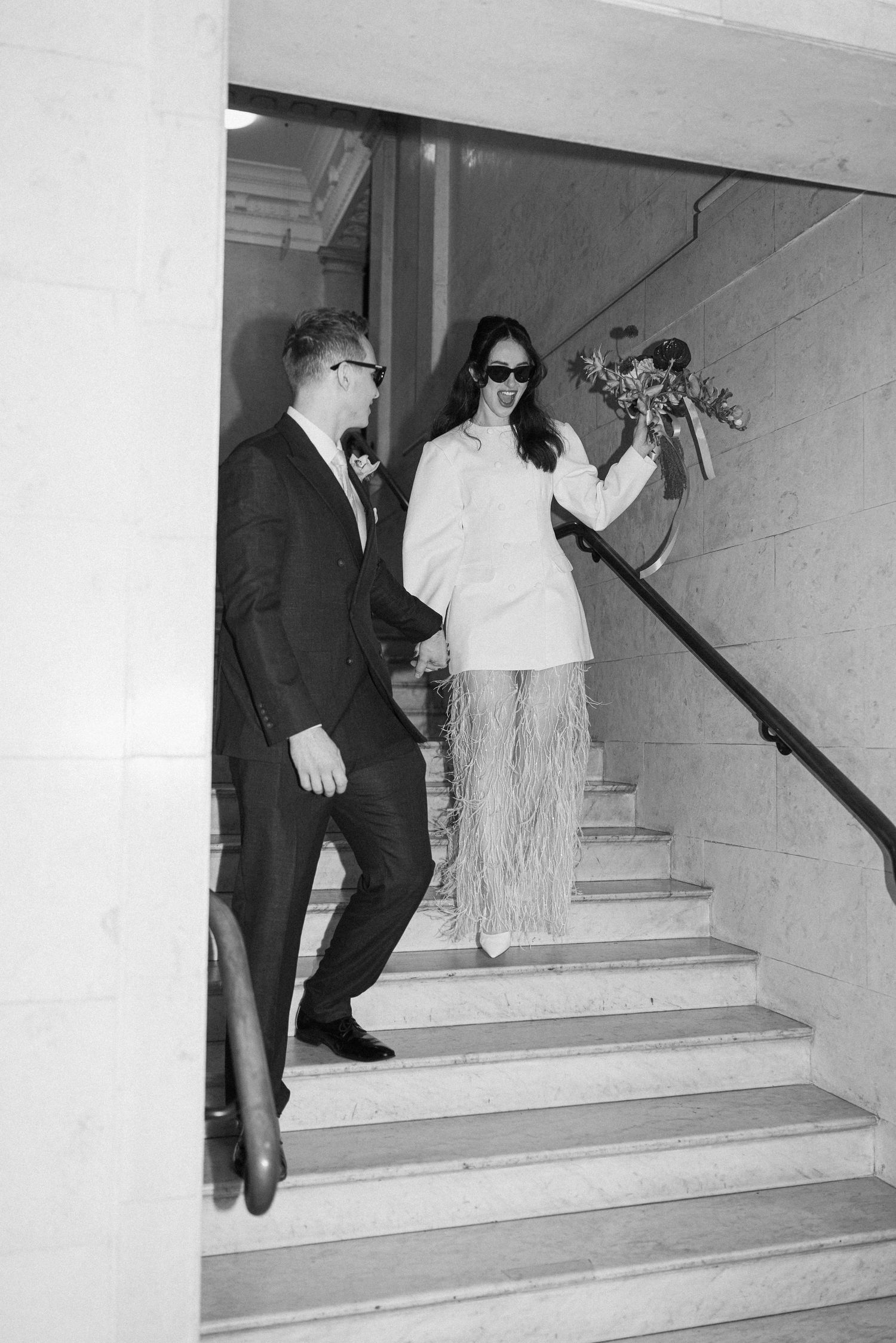 Black and white photo of a bride and groom descending marble stairs holding a floral bouquet.