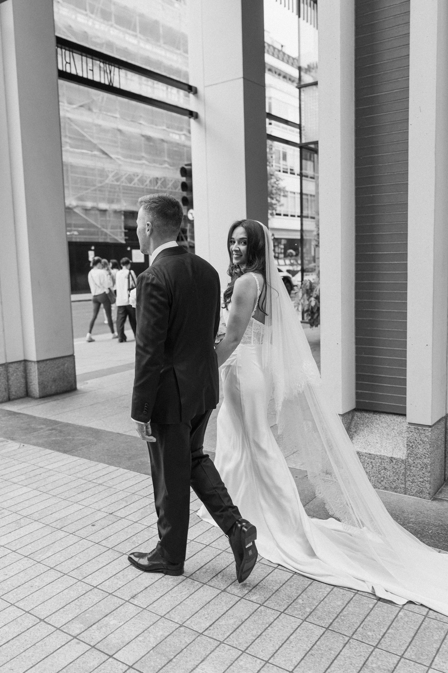 Black and white photo of bride and groom walking hand in hand through an urban city setting.