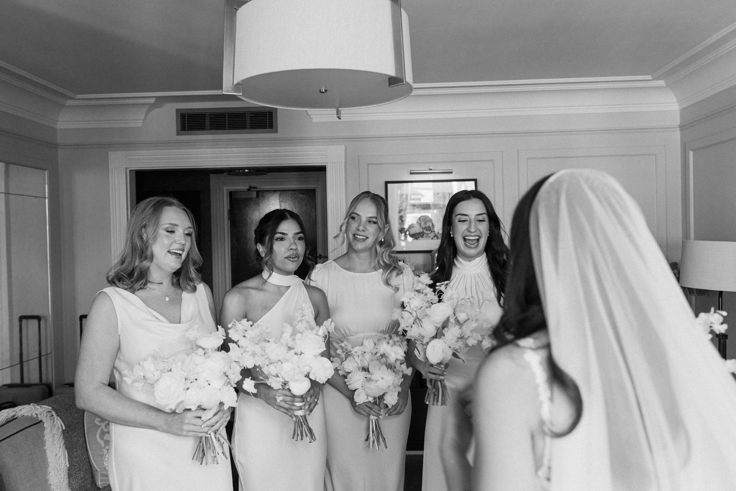Black and white photo of bridesmaids in dresses reacting with joy as veiled bride enters the room.
