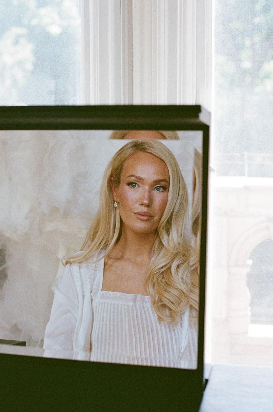 Blonde woman in white top reflected in mirror near bright window, elegant portrait photography.