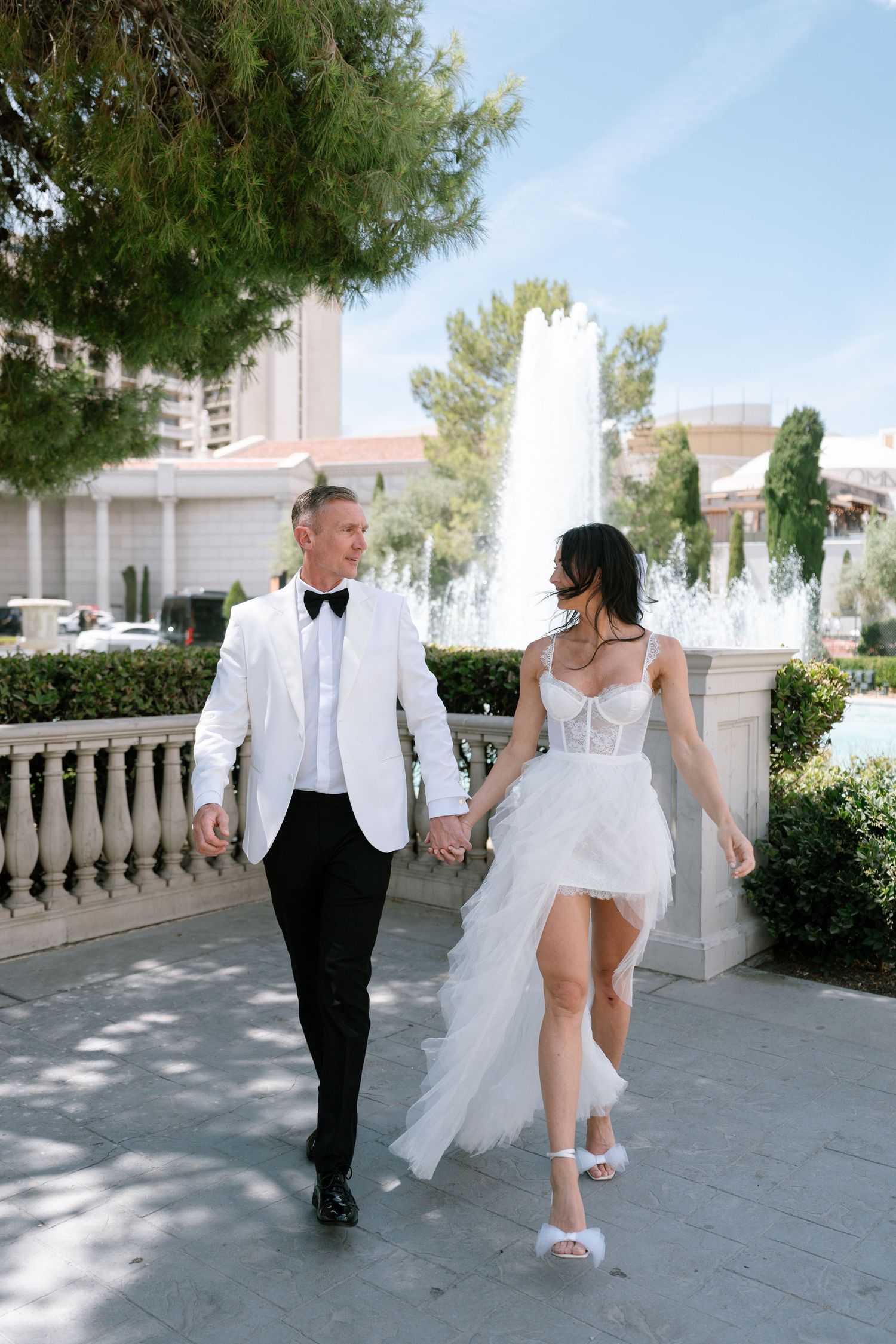 A wedding couple strolls along a path with fountains at a luxury resort, the bride wearing a flowing white dress.