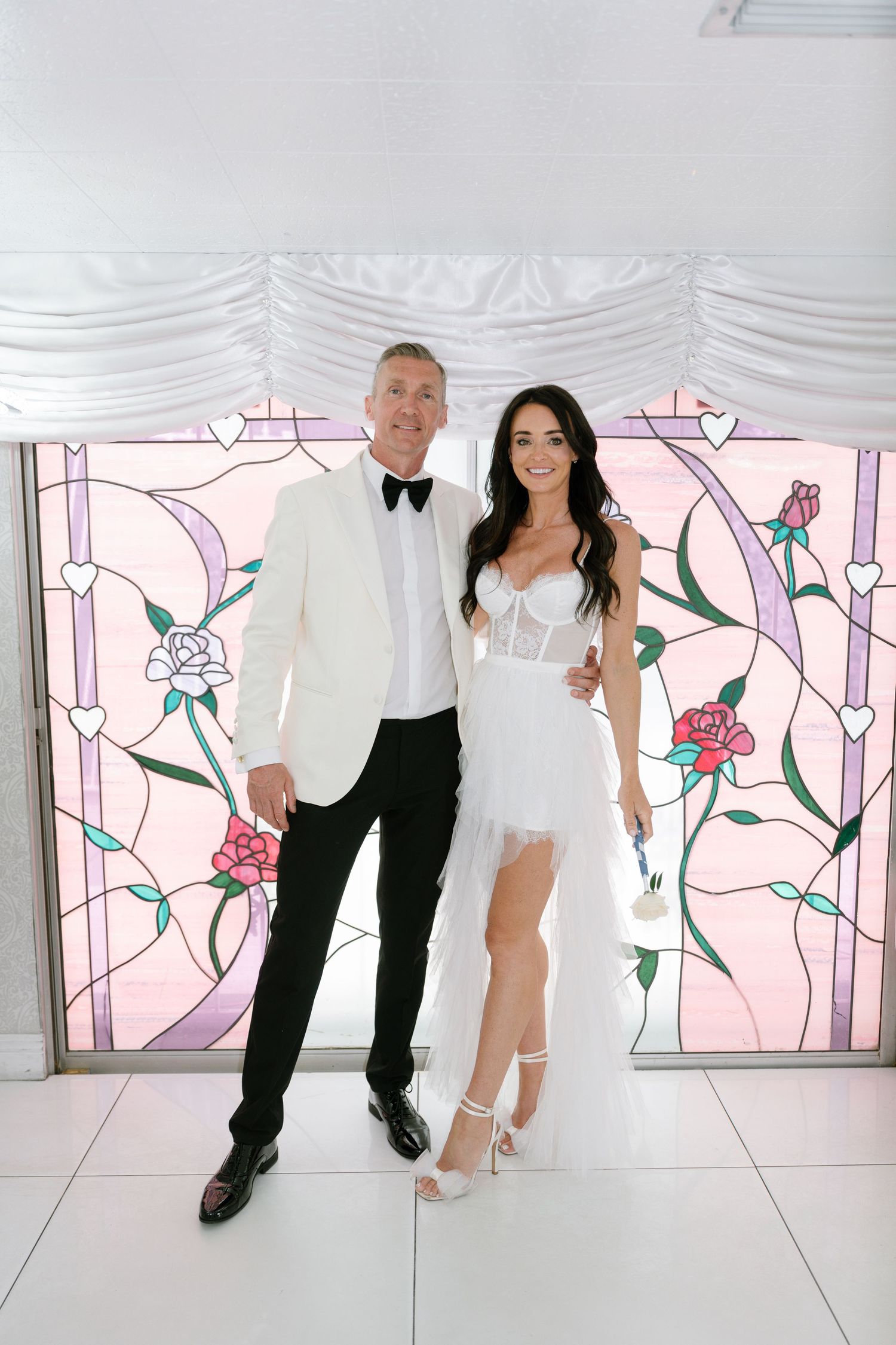 Couple poses playfully against pink floral wall in white wedding attire.