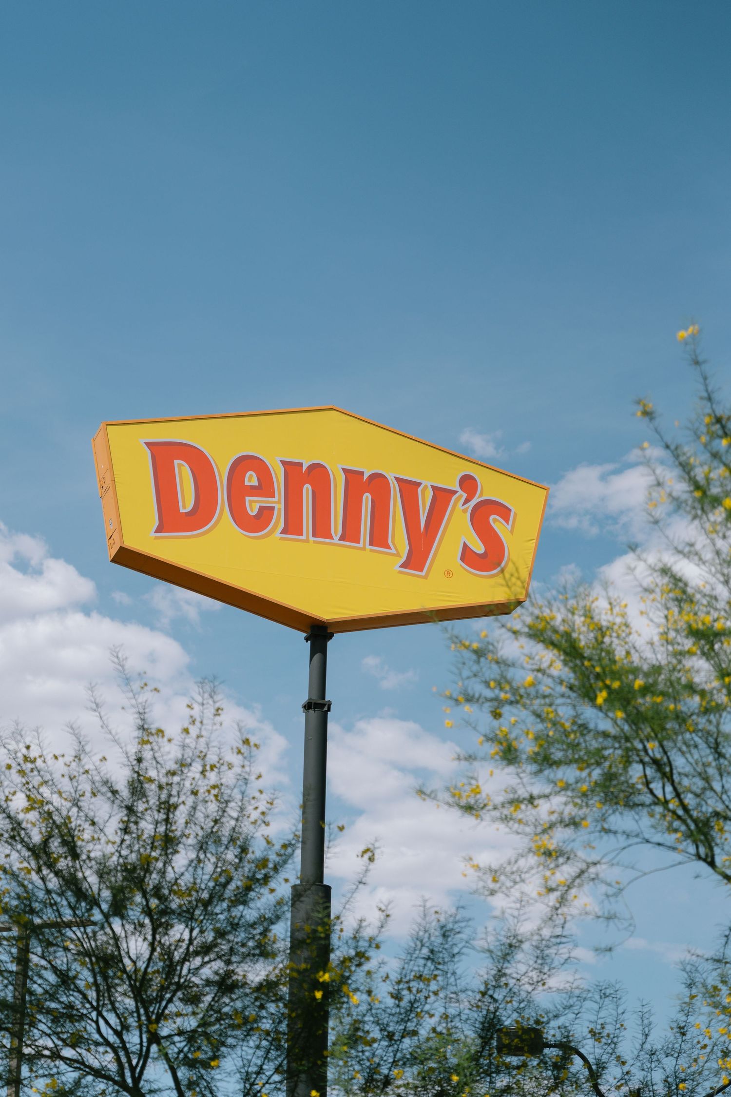 A Denny's restaurant sign against a bright blue sky with white clouds.
