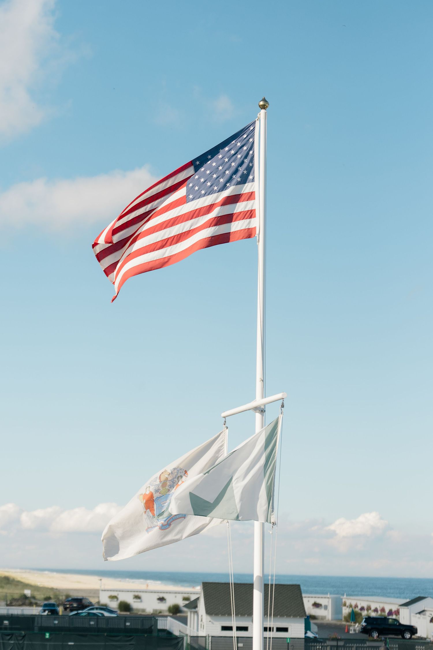 American flag and state flag waving on a white pole with a coastal town and blue sky in the background.