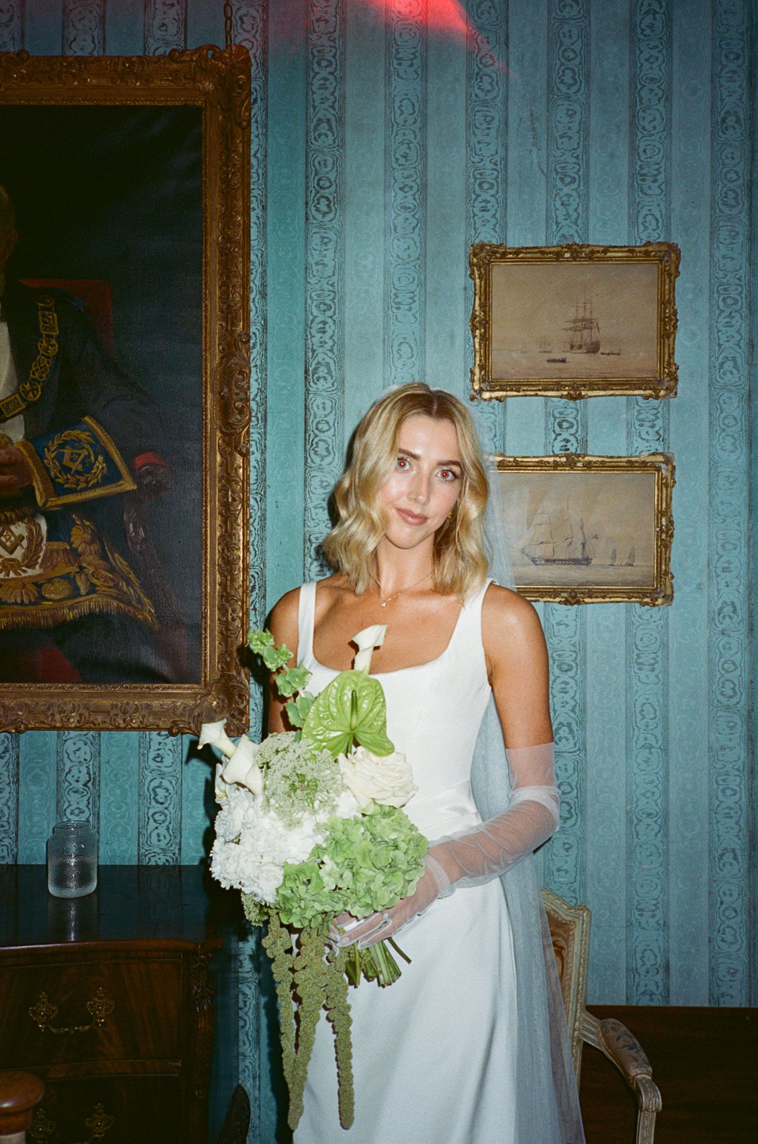 Bride in white gown holding green and white bouquet stands before blue striped walls and ornate gold-framed paintings.