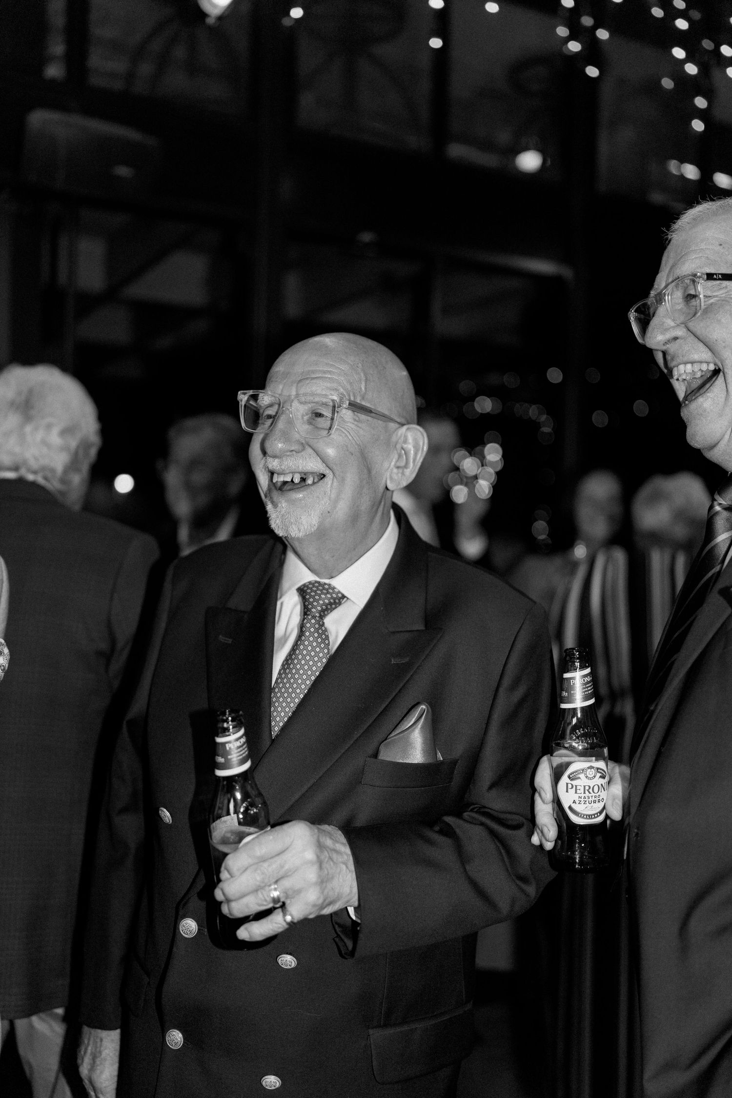 Elderly wedding guest laughing joyfully while holding a beer bottle at an evening wedding reception.
