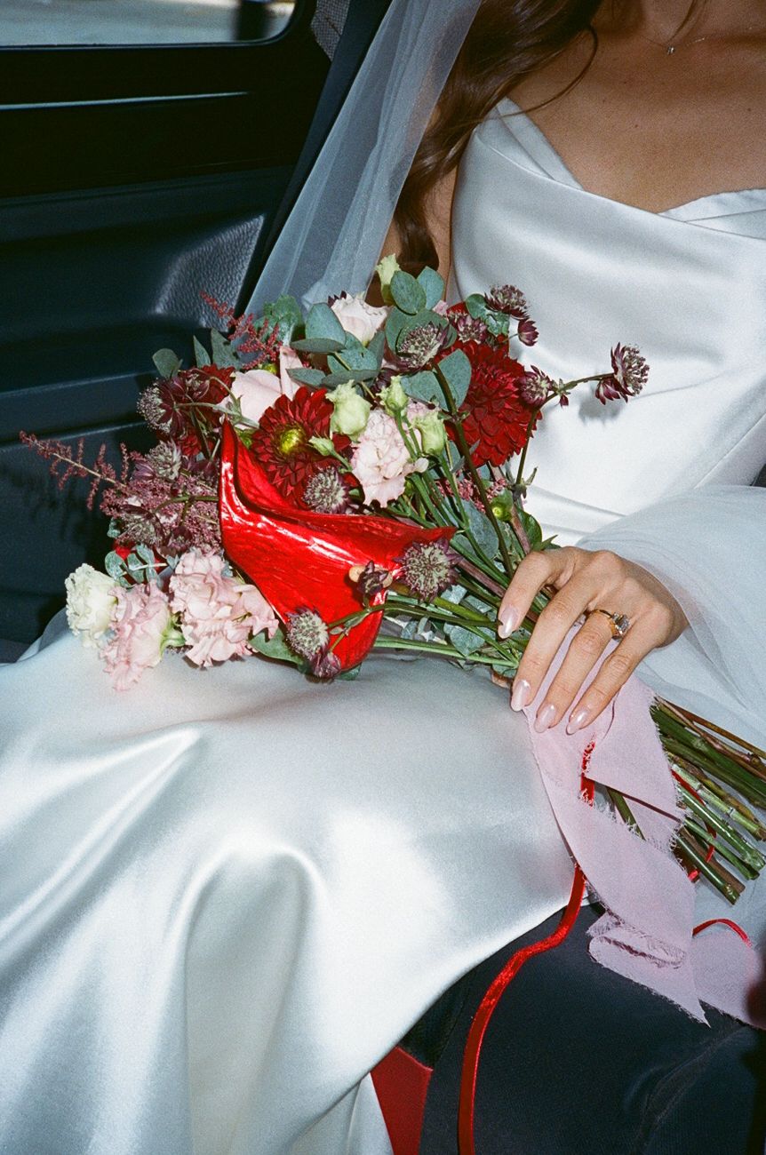 Bride in white satin gown holding red and pink wedding bouquet with cherries in car backseat.