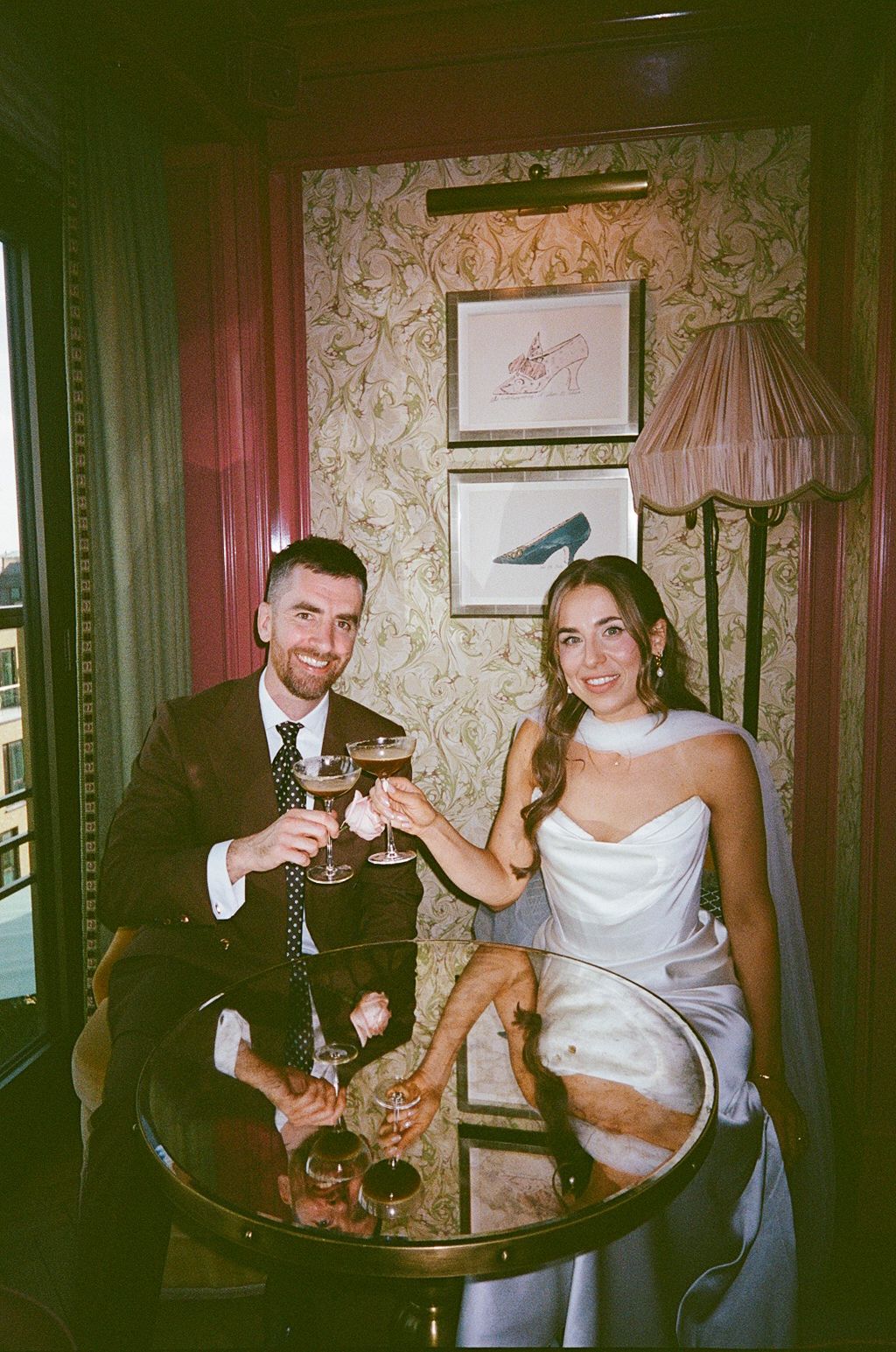 Newlyweds toasting with cocktails at round table in ornate wallpapered venue.