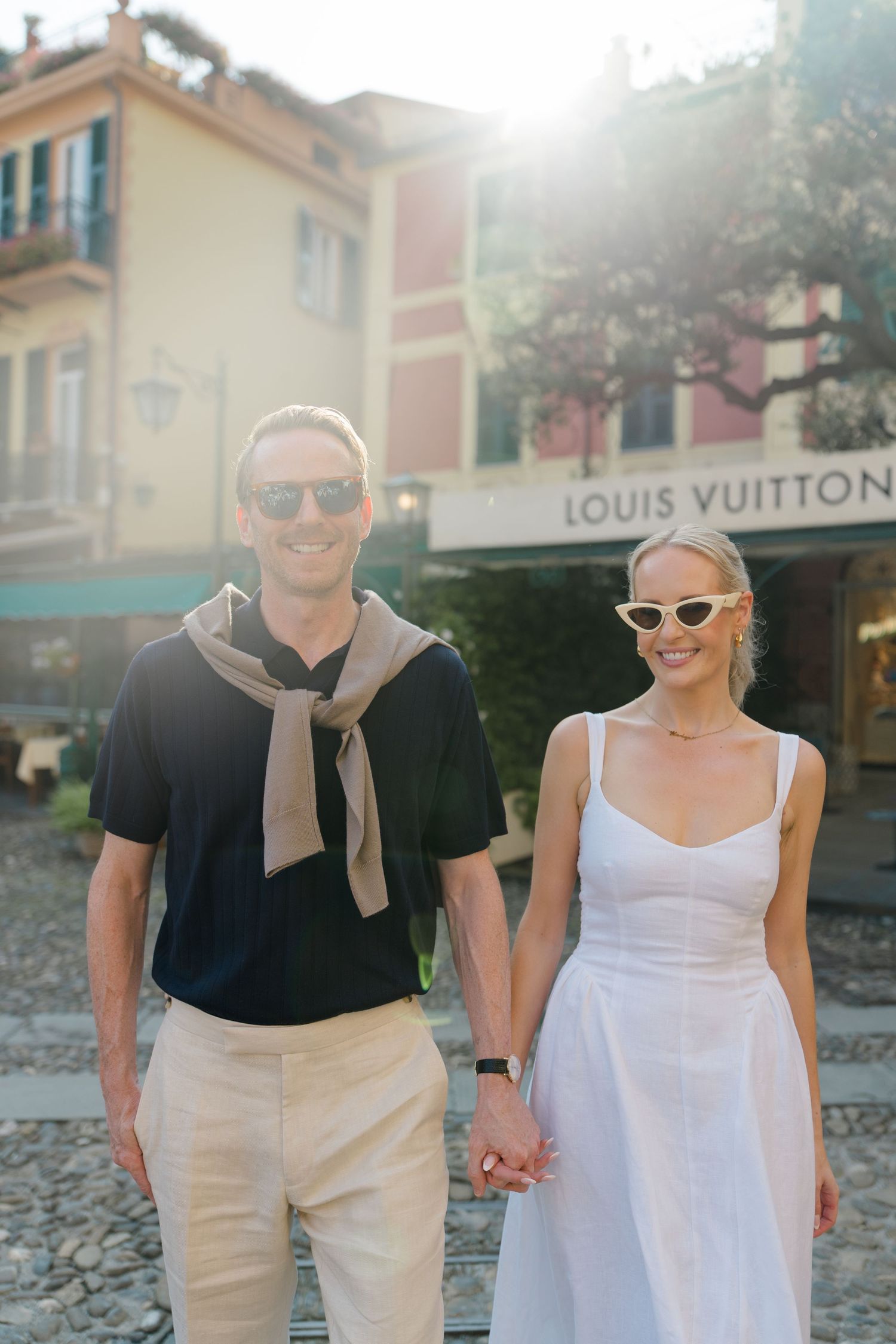 Smiling couple holding hands in front of Louis Vuitton store in Portofino, Italy, wearing sunglasses in golden sunlight.