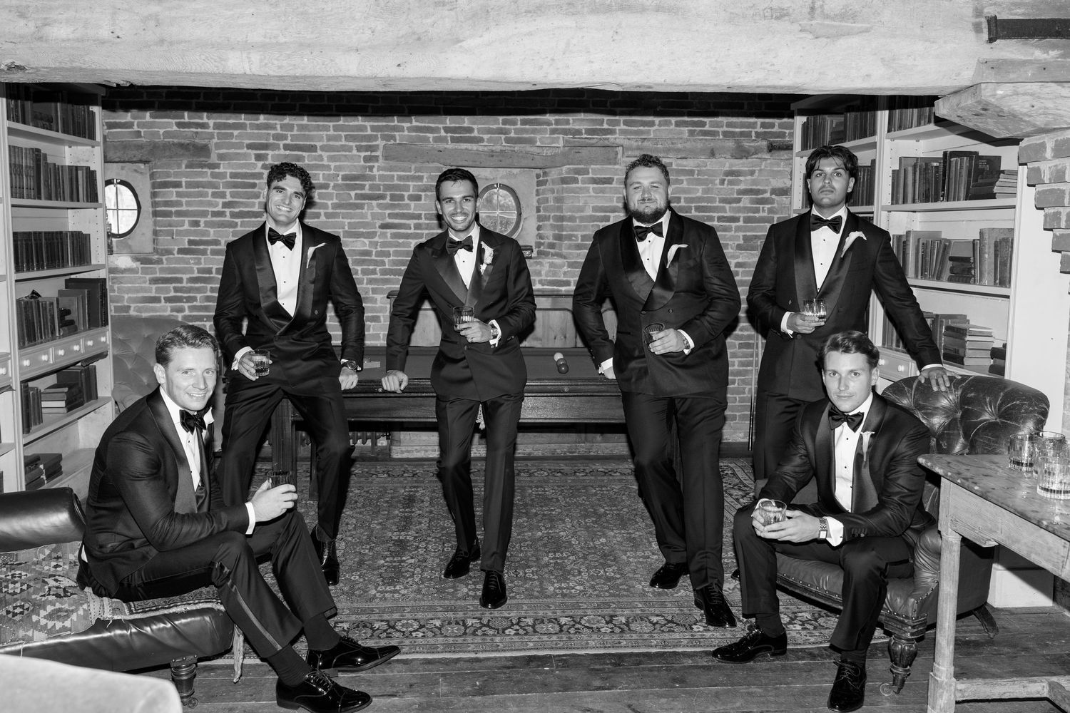 Six groomsmen in black tuxedos pose elegantly in a rustic room with brick fireplace in a black and white photo.