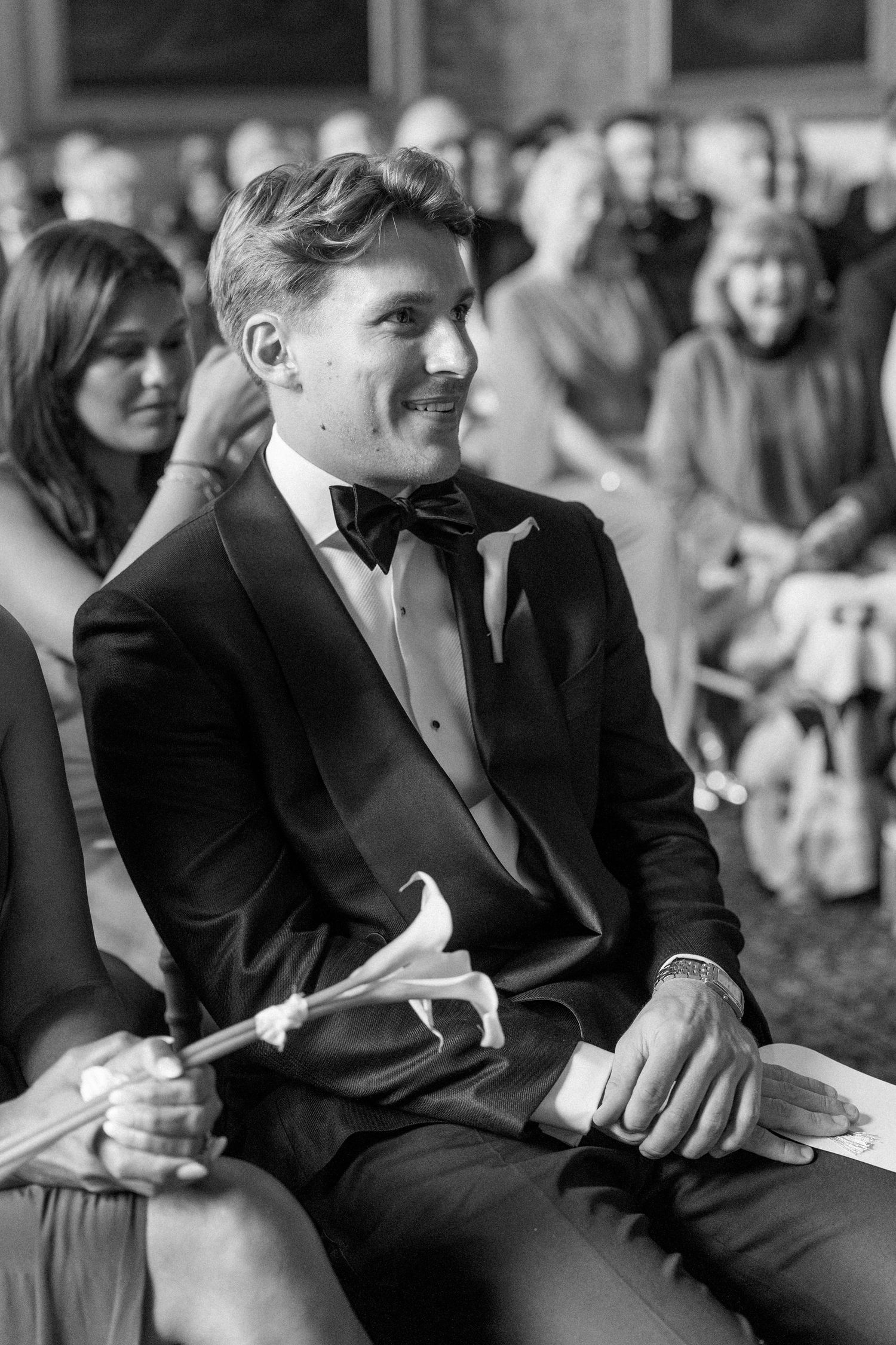 Smiling groom in black tuxedo seated at wedding ceremony holding white flower.