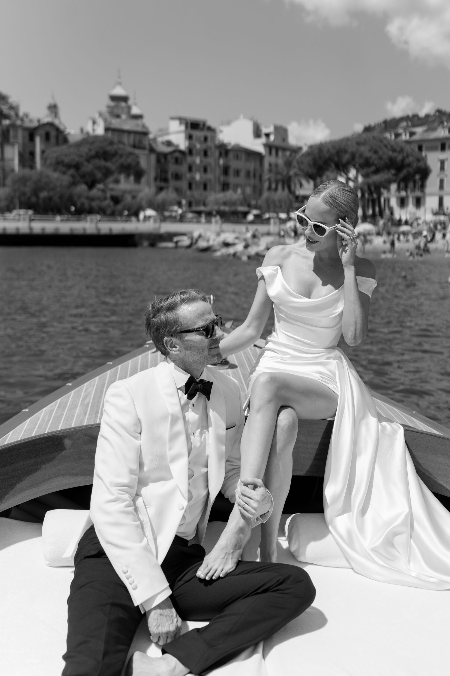 Groom in white tuxedo sits with bride in off-shoulder gown on luxury boat near Italian harbor.