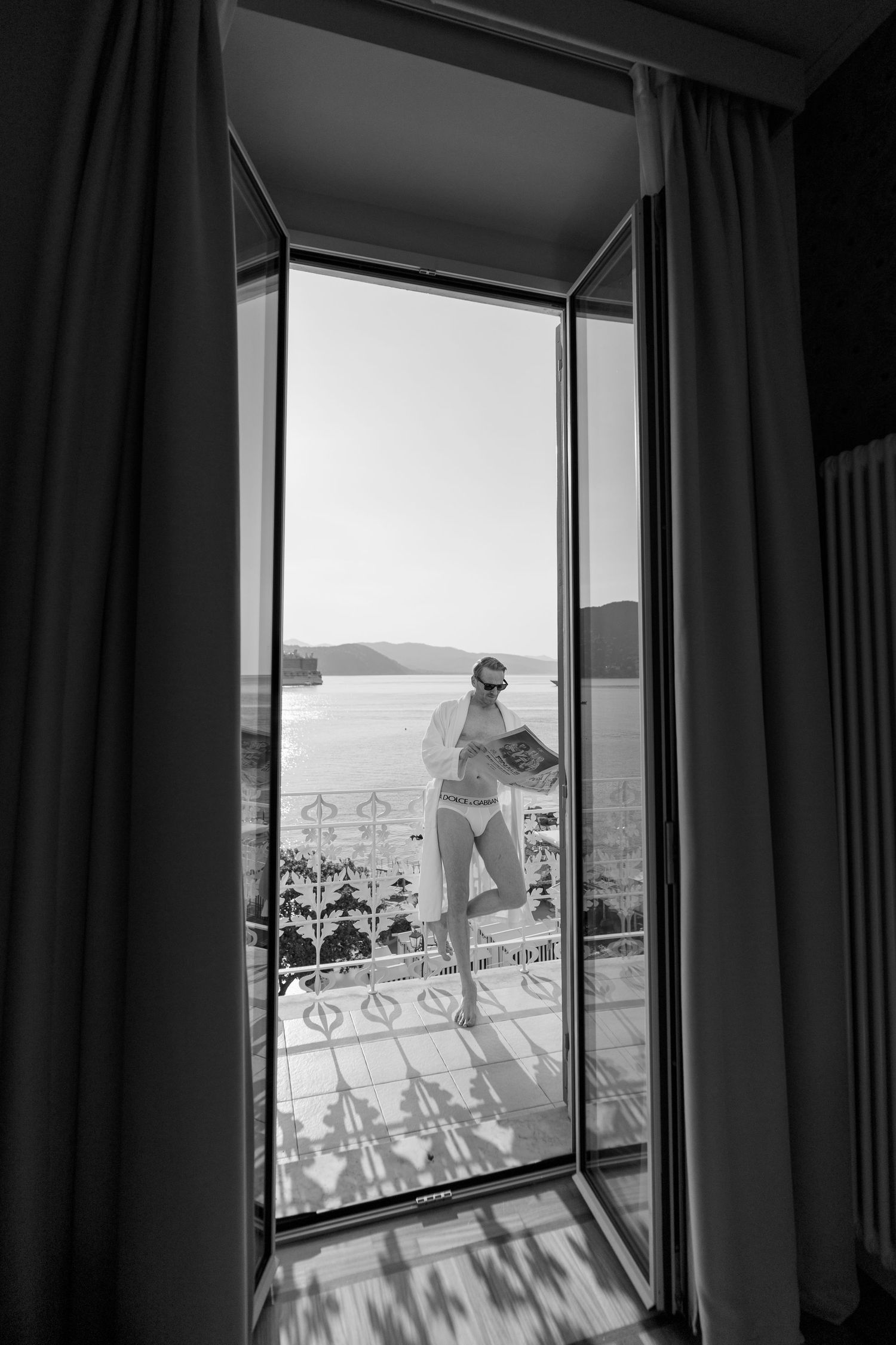 Black and white photo of a woman in bikini standing on a hotel balcony with scenic sea views.