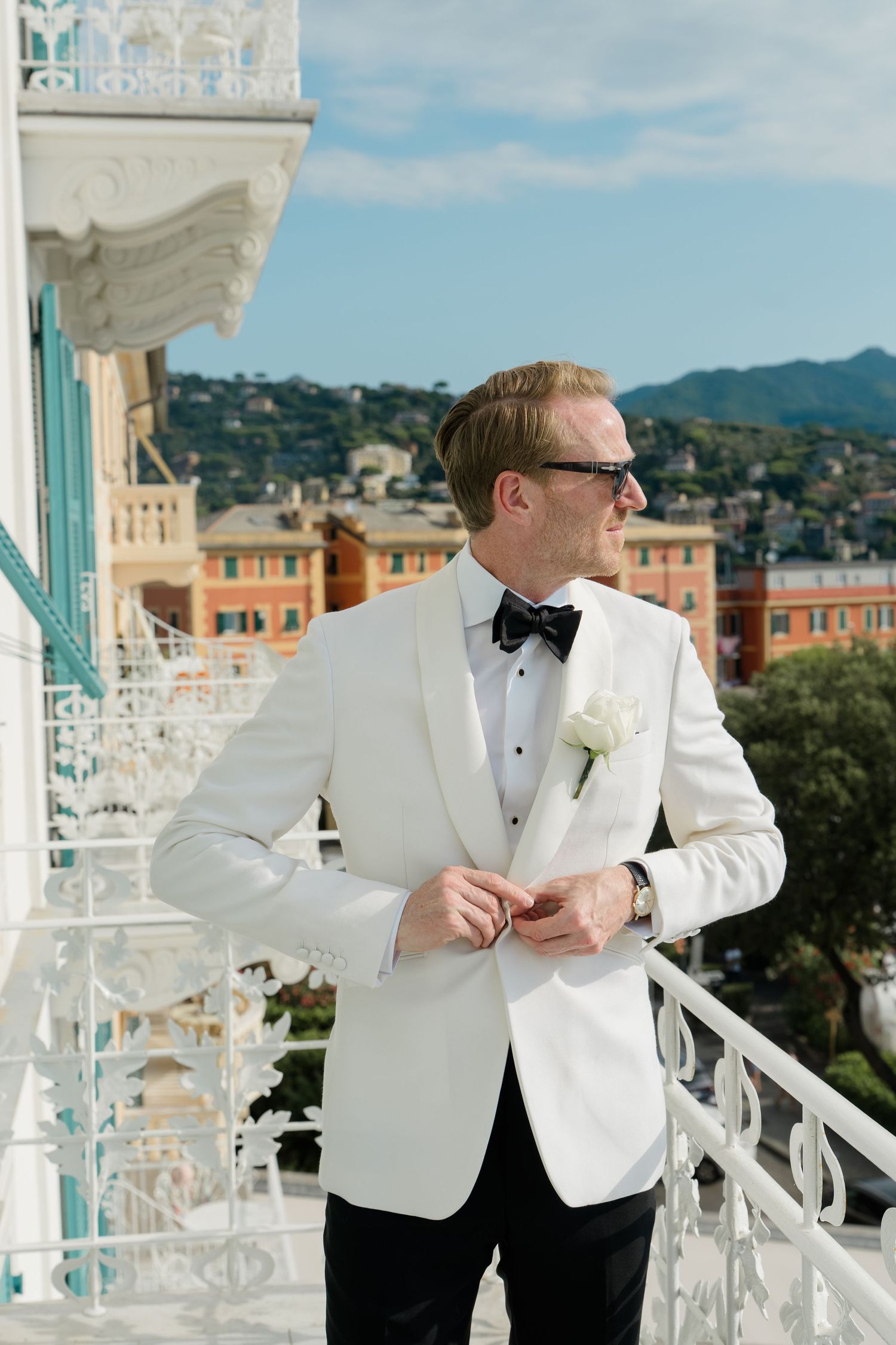 Groom in white tuxedo jacket and bow tie stands on ornate balcony with Italian hillside backdrop.