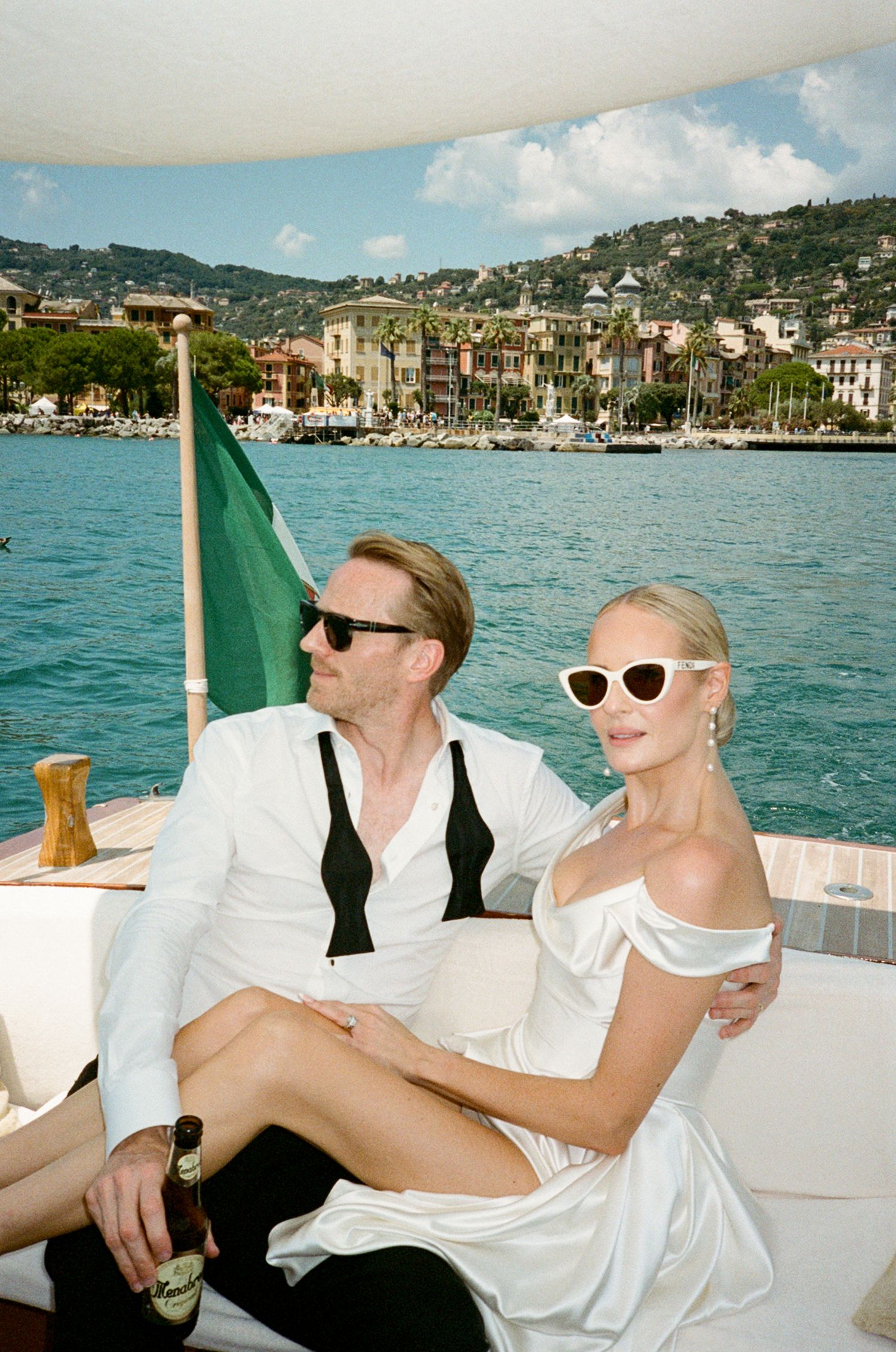 Elegant couple in white attire relaxing on luxury boat with Italian Riviera coastal town backdrop.