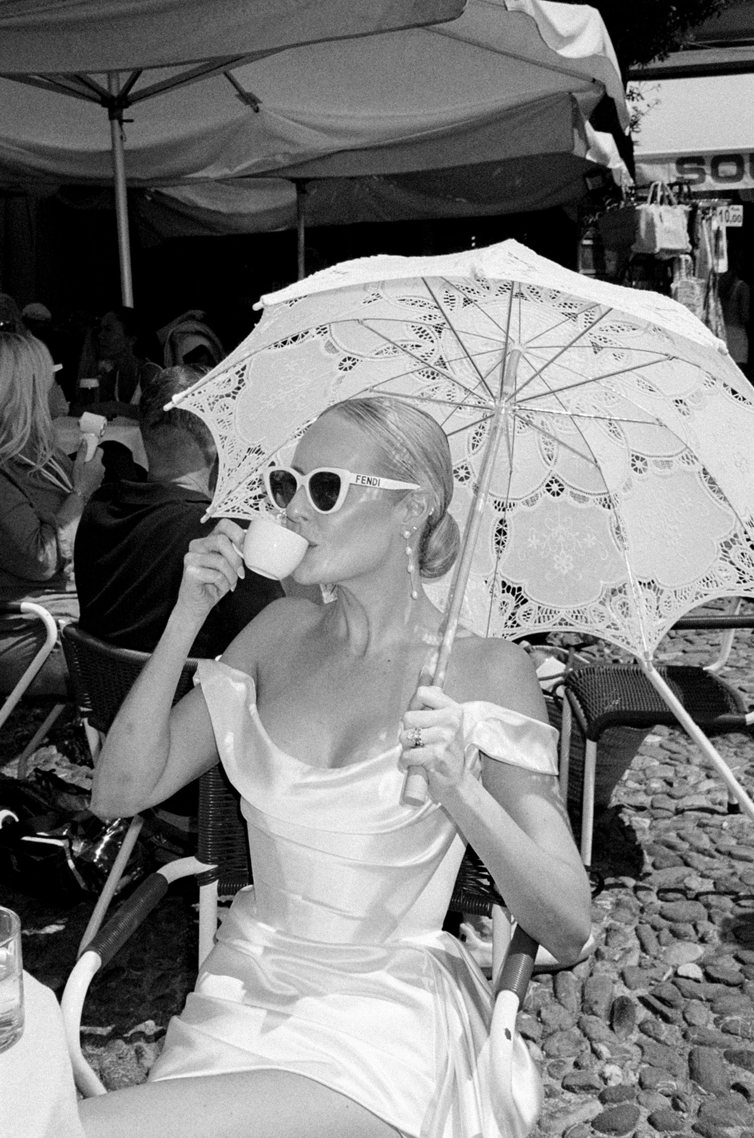 Bride in white gown sips espresso under lace parasol at outdoor cafe, black and white.