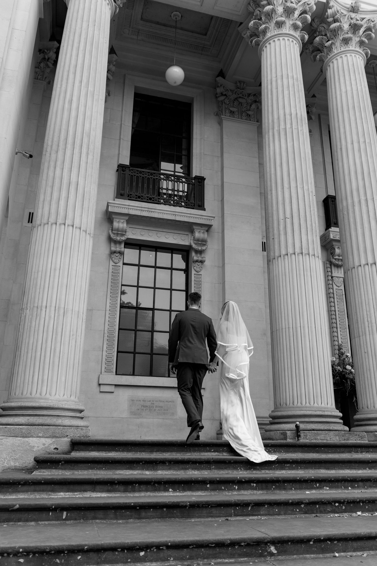 Bride and groom walking up grand stone steps with tall classical columns at a historic wedding venue.