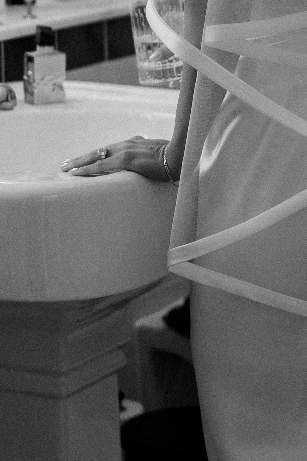 Black and white photo of a bride's hand resting on a pedestal sink, wearing a ring and bracelet in wedding dress.
