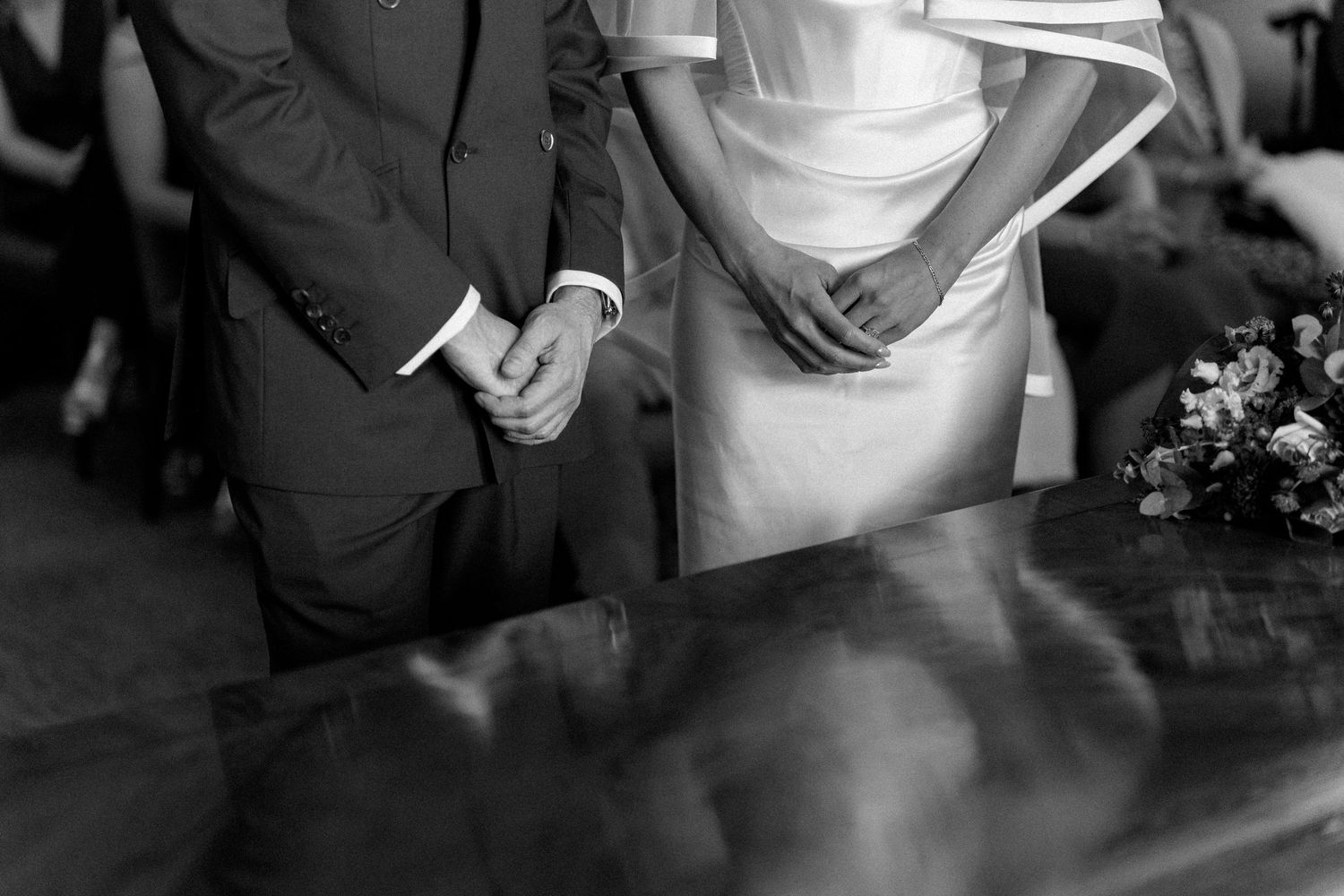 Black and white close-up of bride and groom holding hands at wedding ceremony register table.