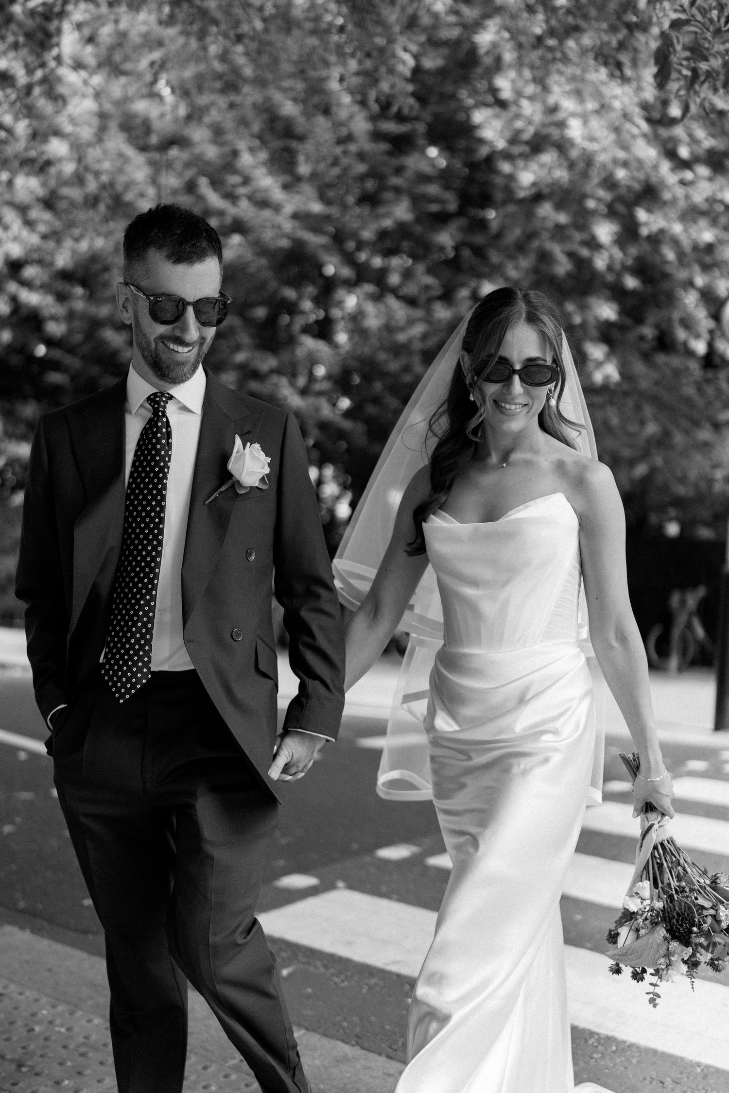 Black and white photo of smiling bride and groom in sunglasses walking hand in hand outdoors.