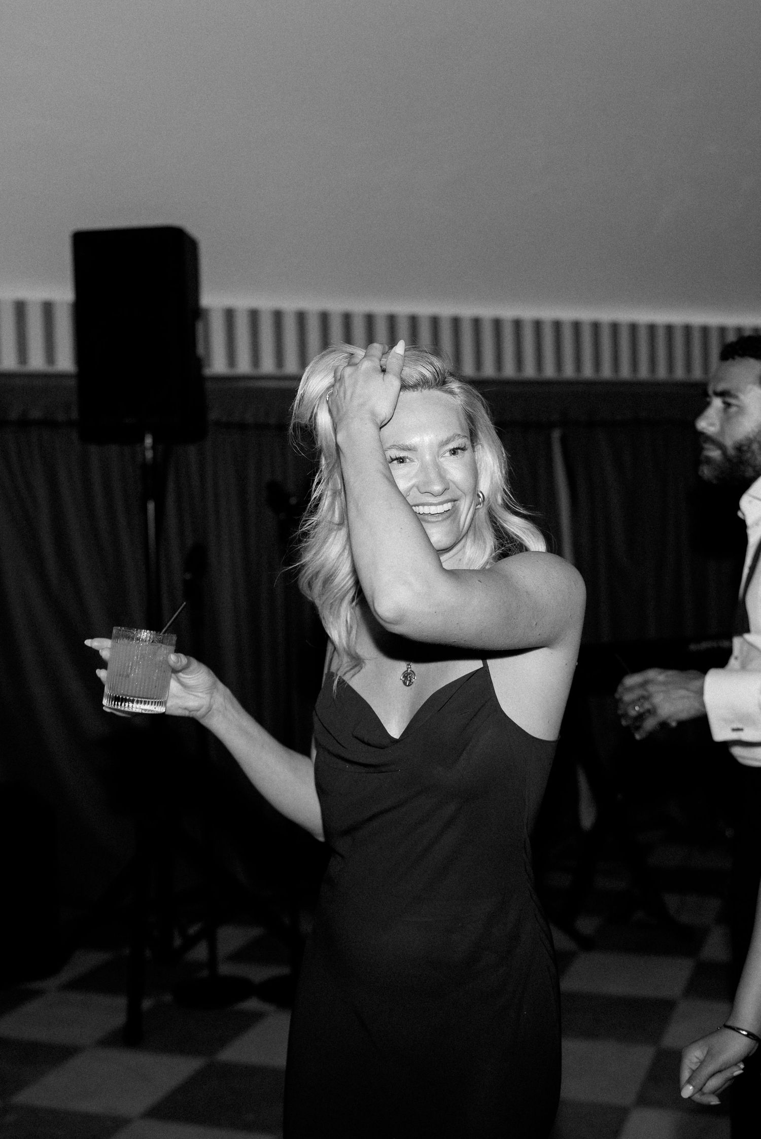 Woman in black dress laughing and dancing with a cocktail at a wedding reception party.