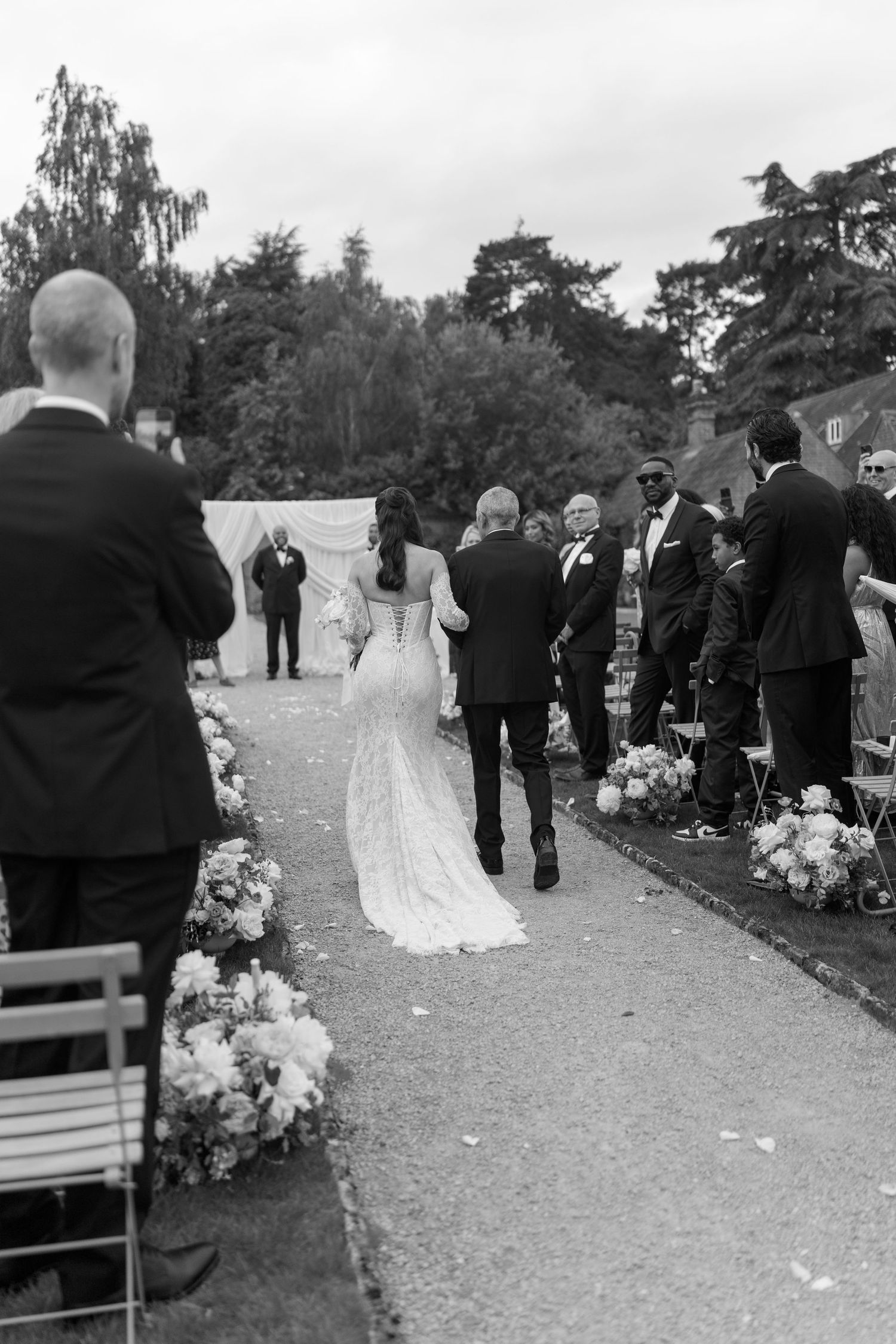 Bride in lace gown walking down a flower-lined outdoor aisle with her father at a garden wedding.