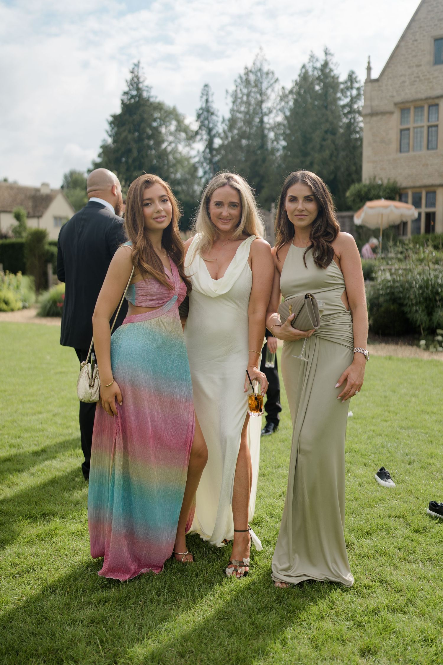Three women in elegant gowns pose together on lawn outside stone manor at summer wedding.