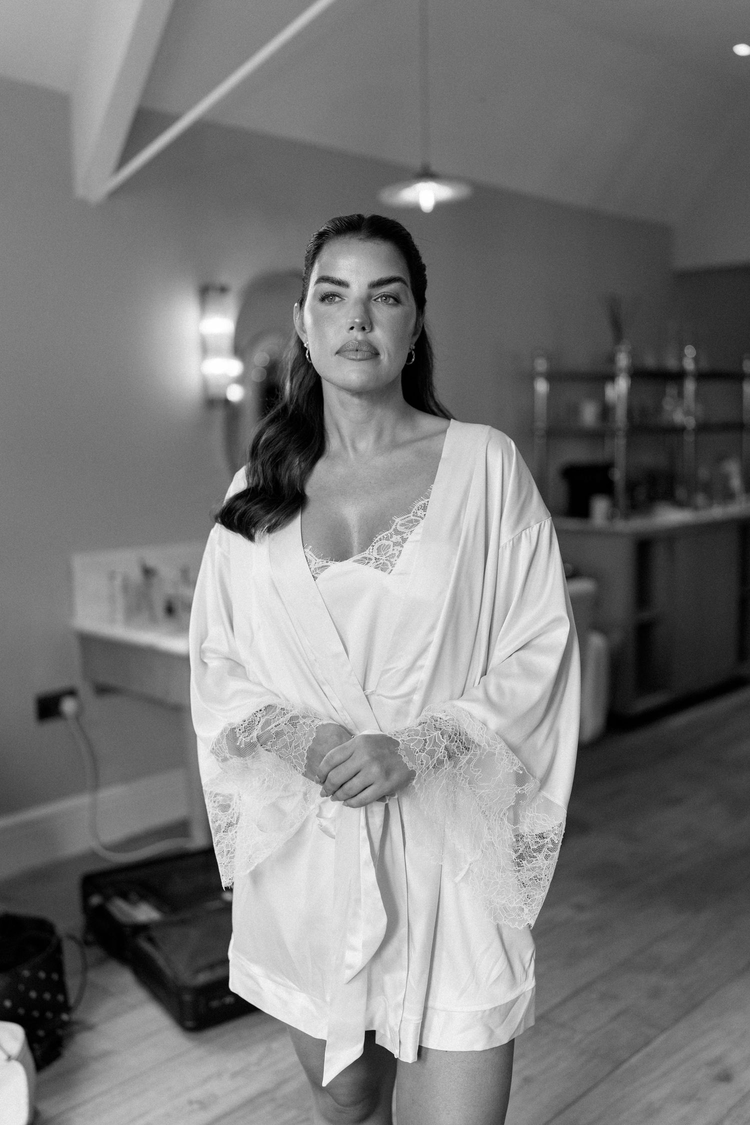 Black and white portrait of a bride in a white lace robe standing in a bridal suite.