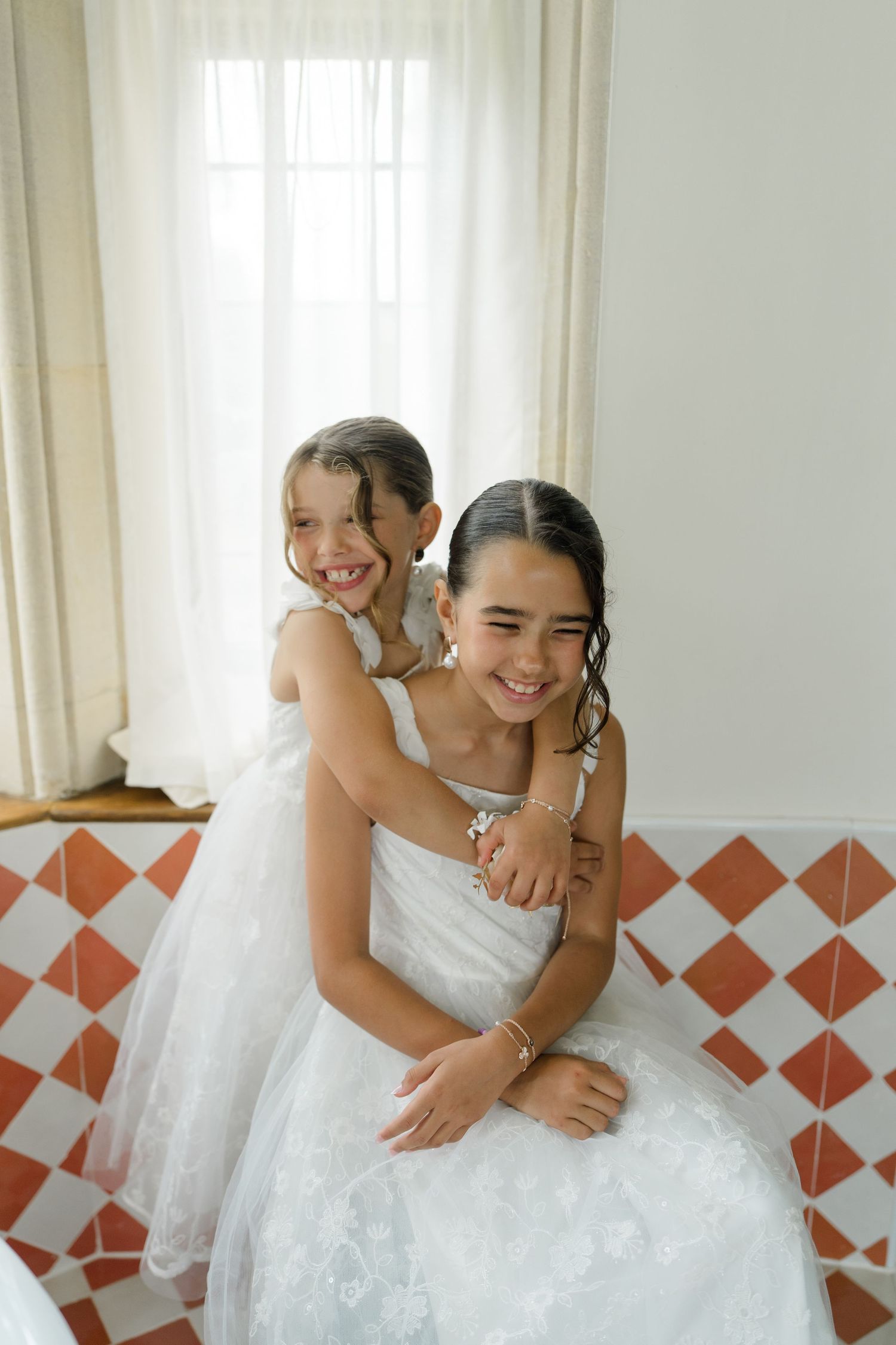Two flower girls in white dresses hugging and smiling by a checkered tile window seat.